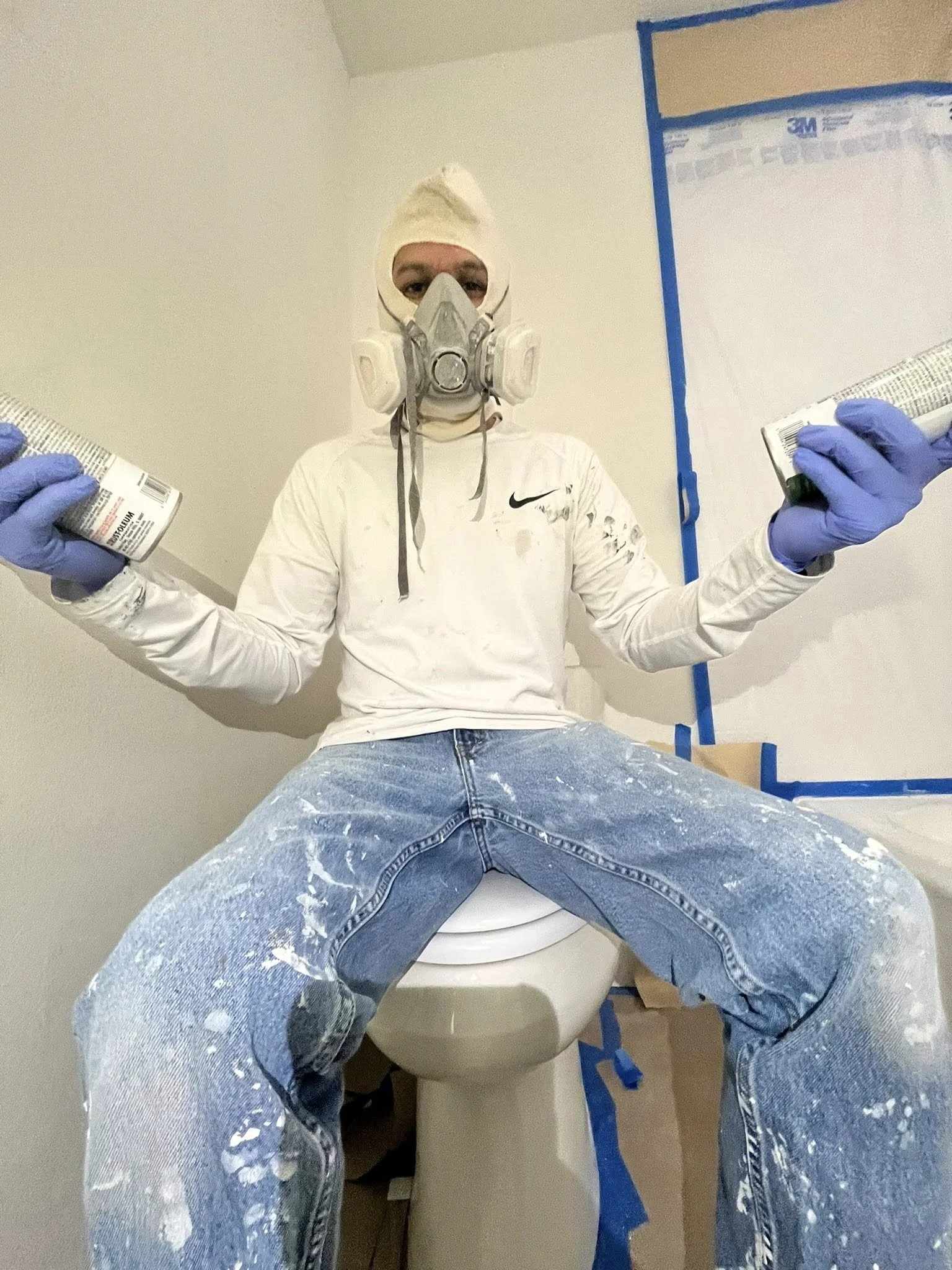 A person sitting on a toilet seat wearing a mask, goggles, gloves, and a hoodie, holding spray cans, in a room under renovation.