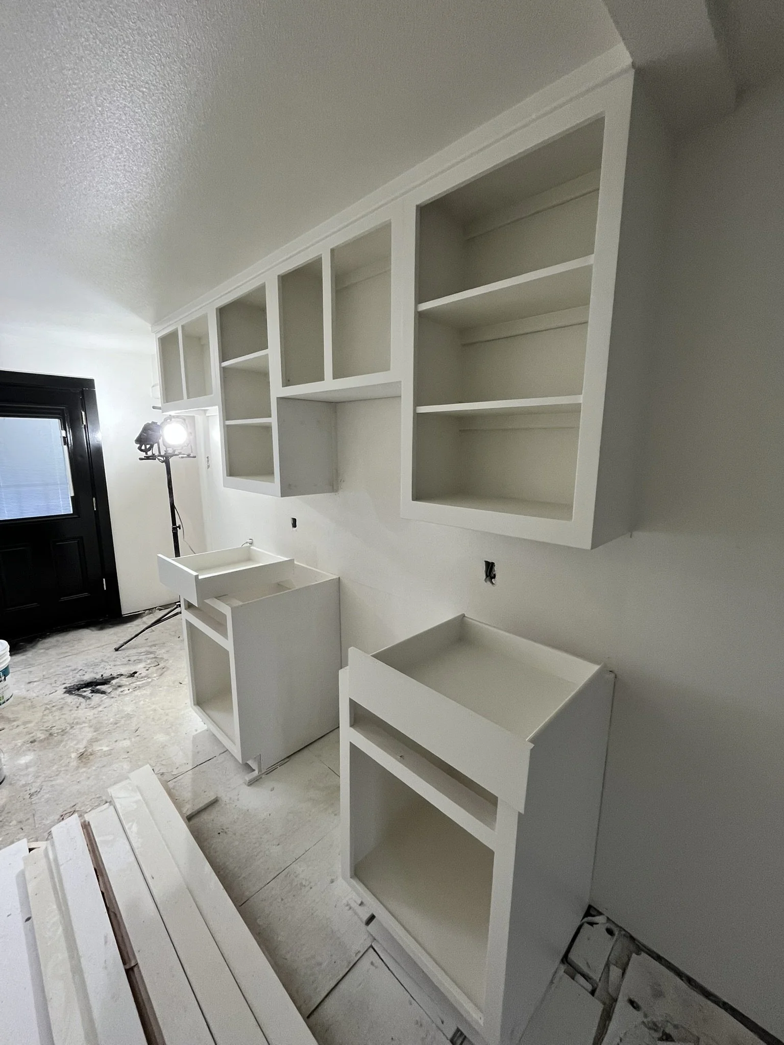 White kitchen cabinets being installed on a wall, with some upper cabinets open and no doors, and two lower cabinet frames without doors.