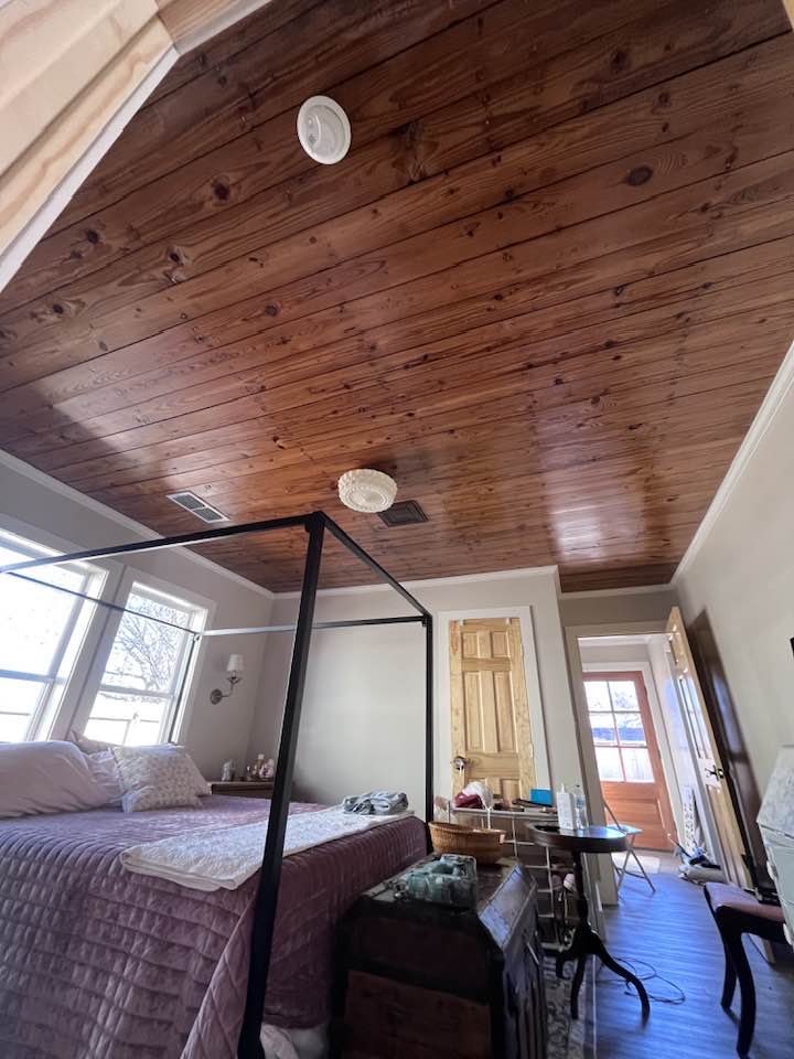 A bedroom with a wooden ceiling, a bed with a quilt, a window, a small table with water bottles, a chair, and an open door showing a porch outside.