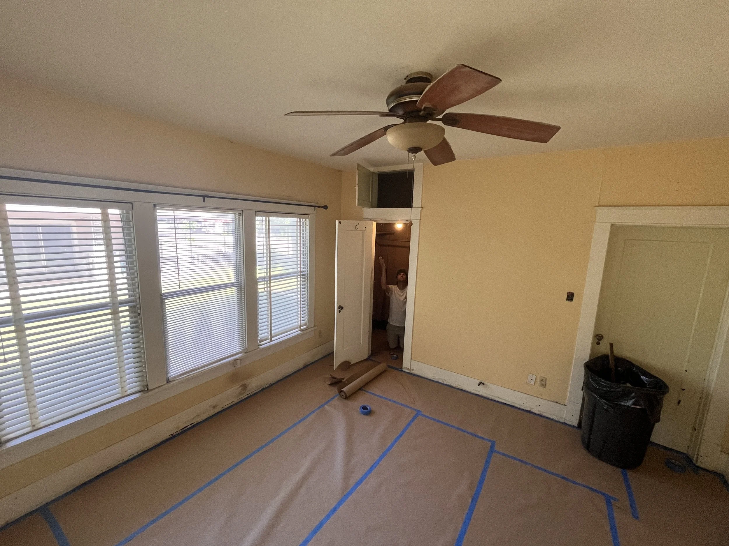 Room undergoing renovation with large windows with blinds, a ceiling fan, and walls painted yellow, with baseboards covered for painting, and tools on the floor.