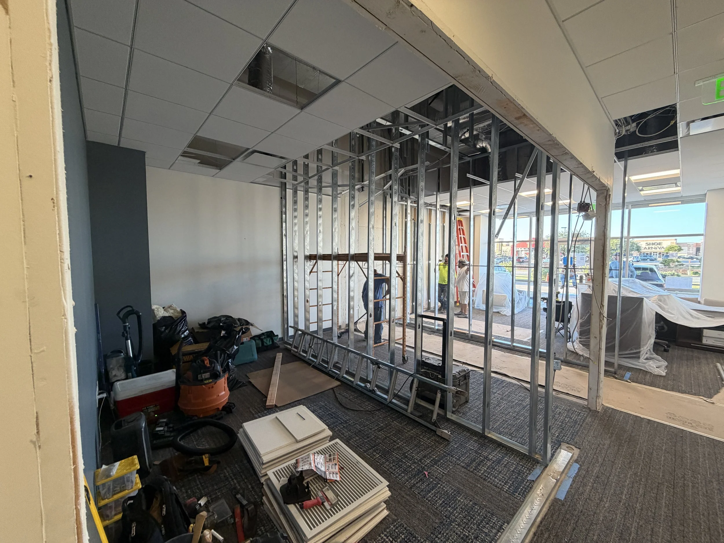The image shows an indoor construction site with framed walls and suspended ceiling panels being installed. Tools and construction materials are scattered on the floor, and workers are in the background.