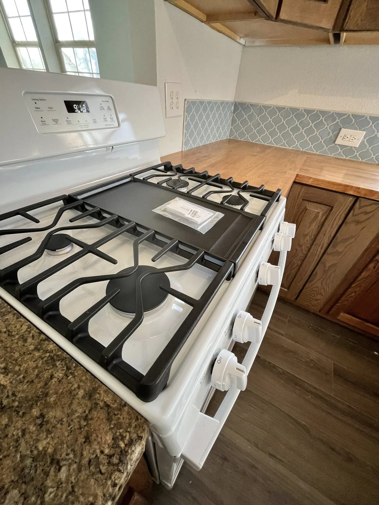 White gas stove with black burners in a kitchen with granite countertop and wooden cabinets, blue patterned backsplash, and electric outlets on the wall.