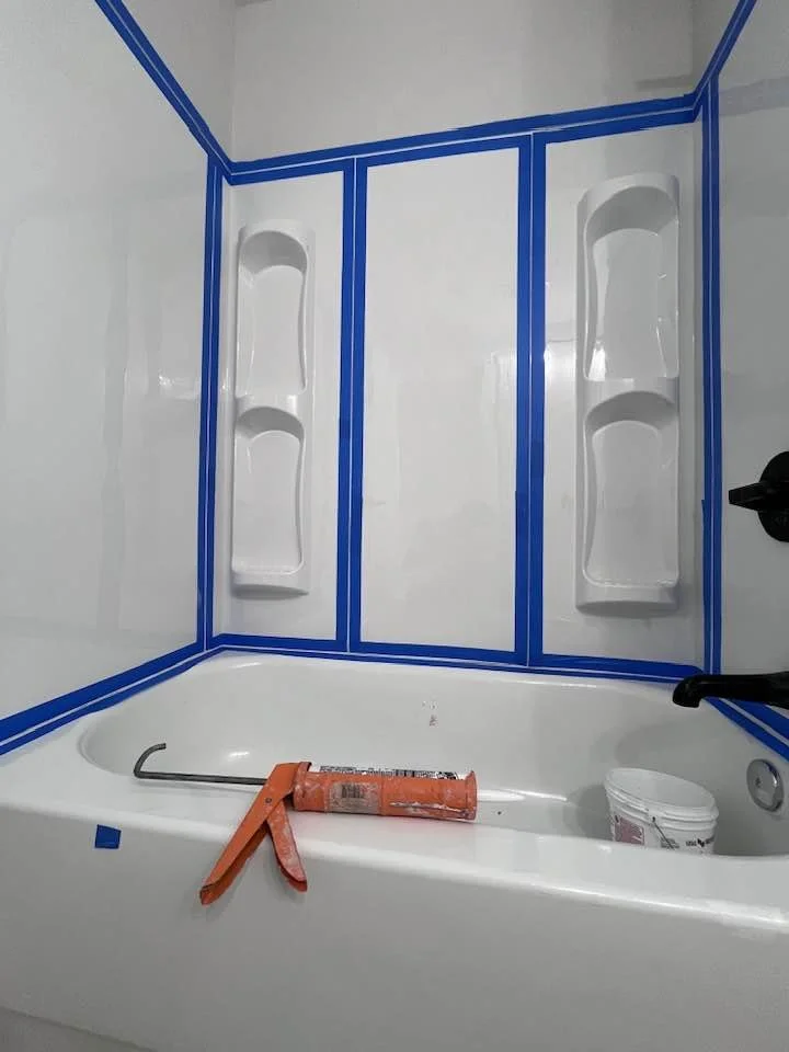 Bathroom shower area with white walls, built-in shelves on either side, and a bathtub with a caulking gun, bucket, and black handle at the edge.