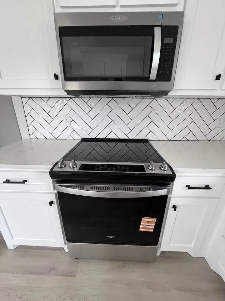 Kitchen with a black and silver electric stove and a microwave oven above it, white cabinets, and a white tile backsplash with a herringbone pattern.