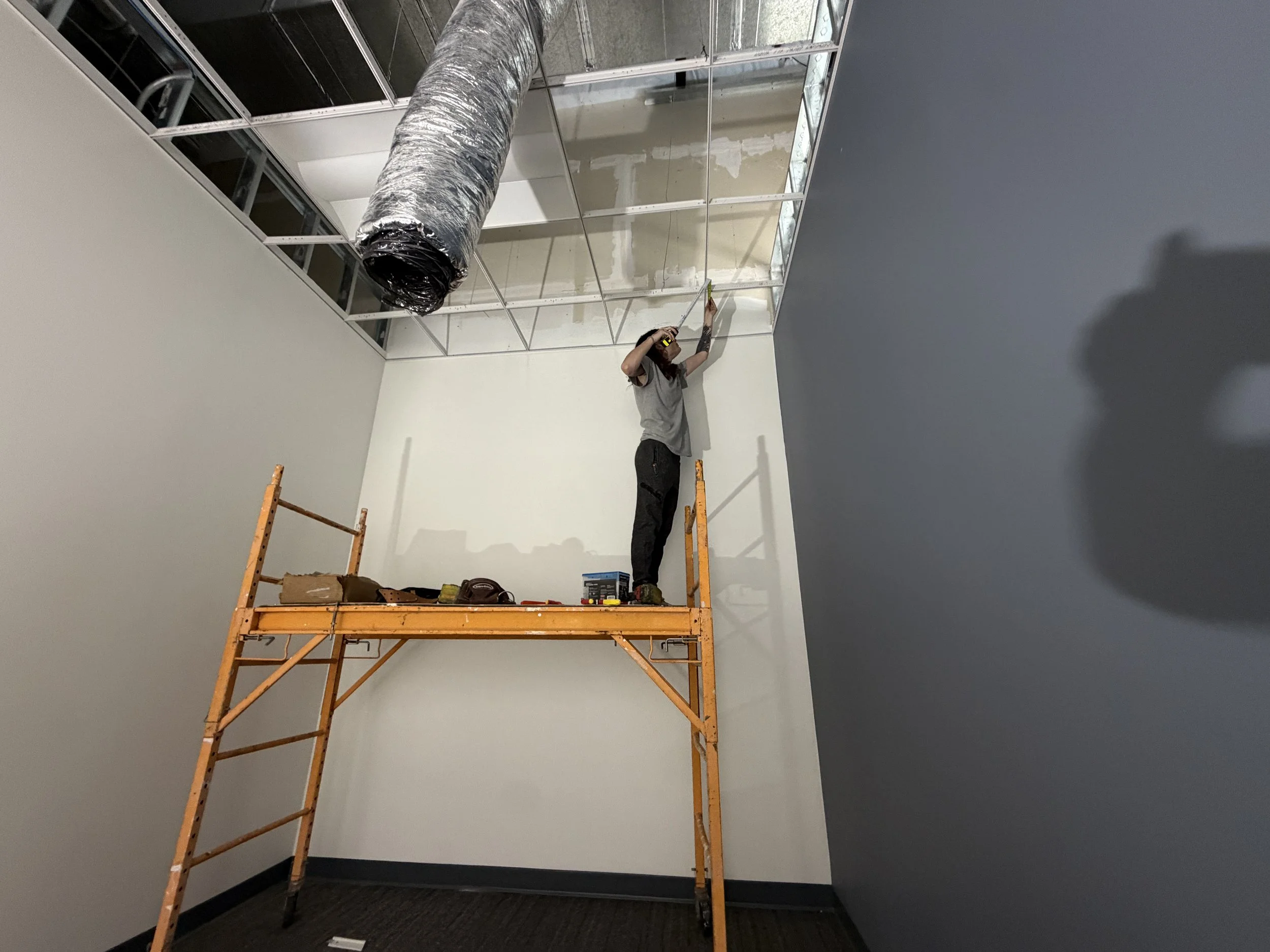 A person standing on an orange scaffolding, installing or working on dry wall behind metal framing in a room under construction. The person is using a tool and is surrounded by construction materials.