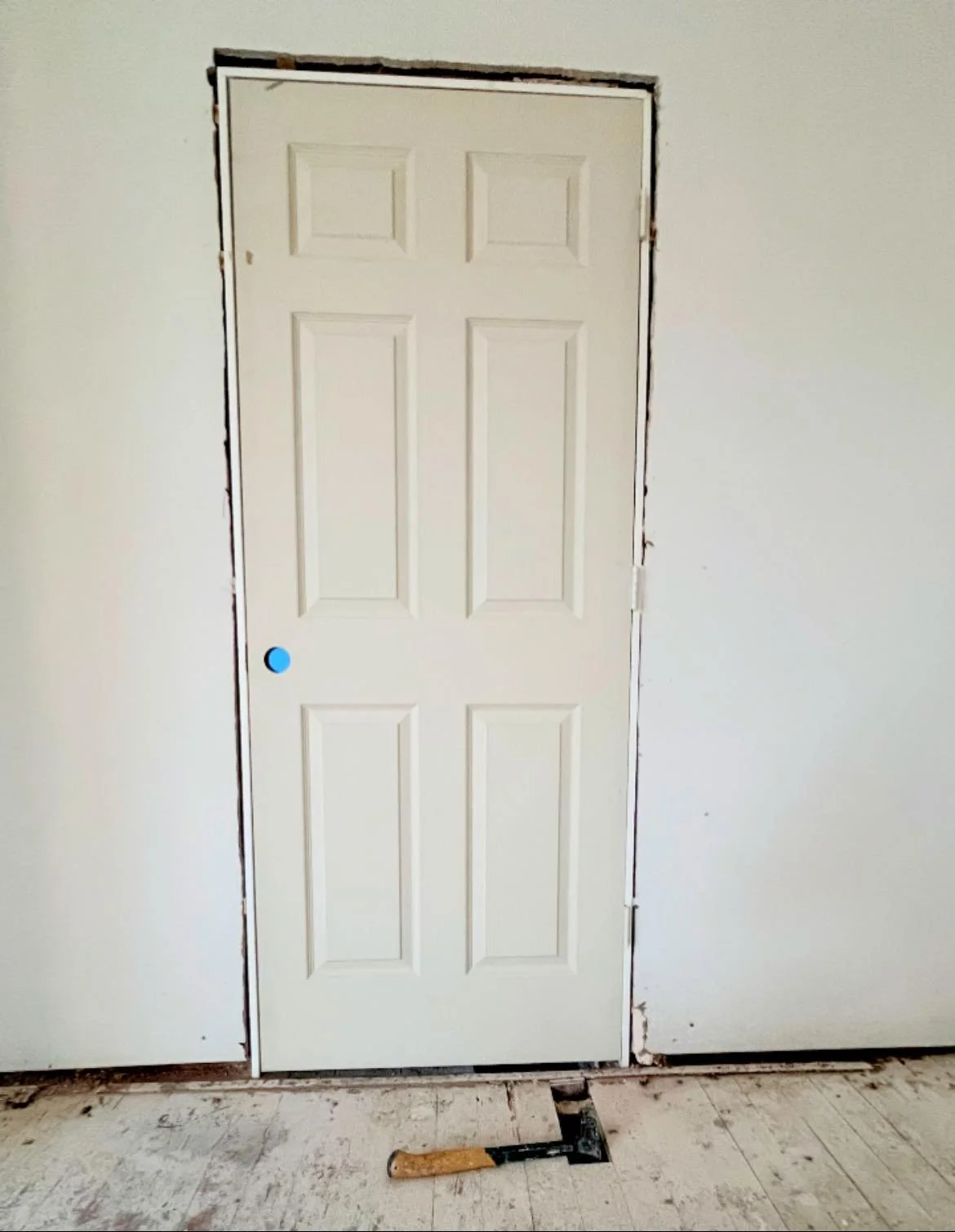 Interior door with unfinished edges and a blue dot near the knob, with construction tools and exposed floor underneath.