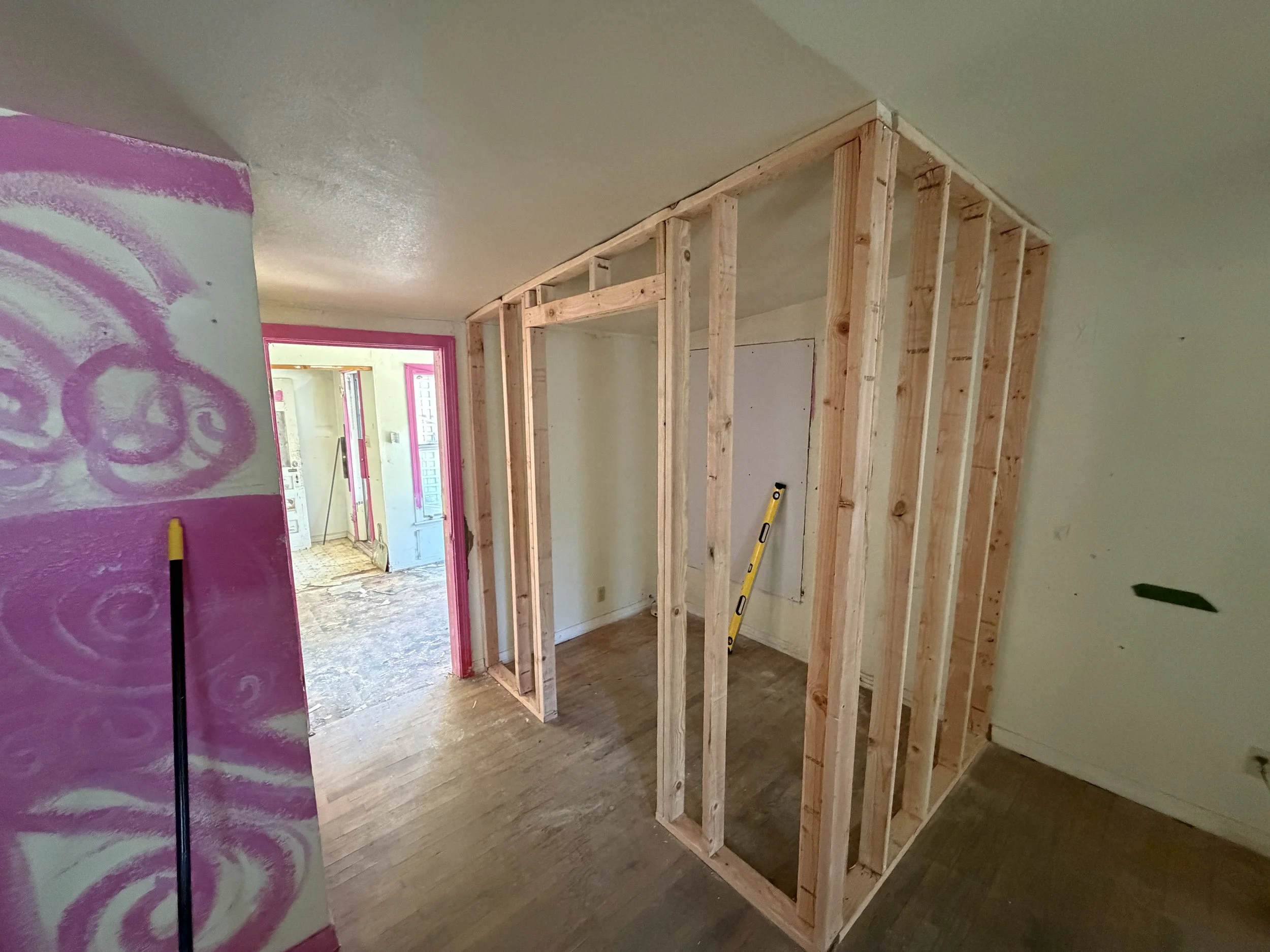Indoor construction site with a wooden wall frame, partially painted pink wall on the left, and an open doorway leading to a brightly lit area.