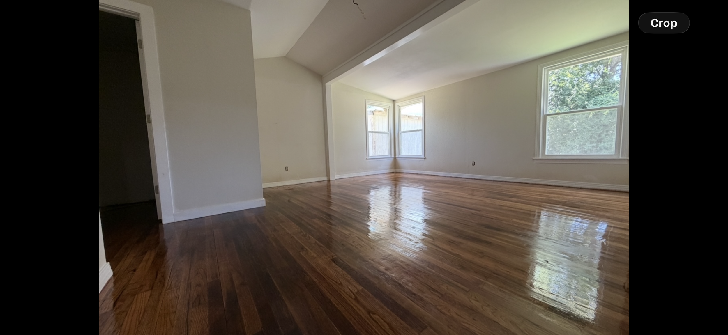 Empty living room with hardwood floors, white walls, and large windows showing green trees outside.
