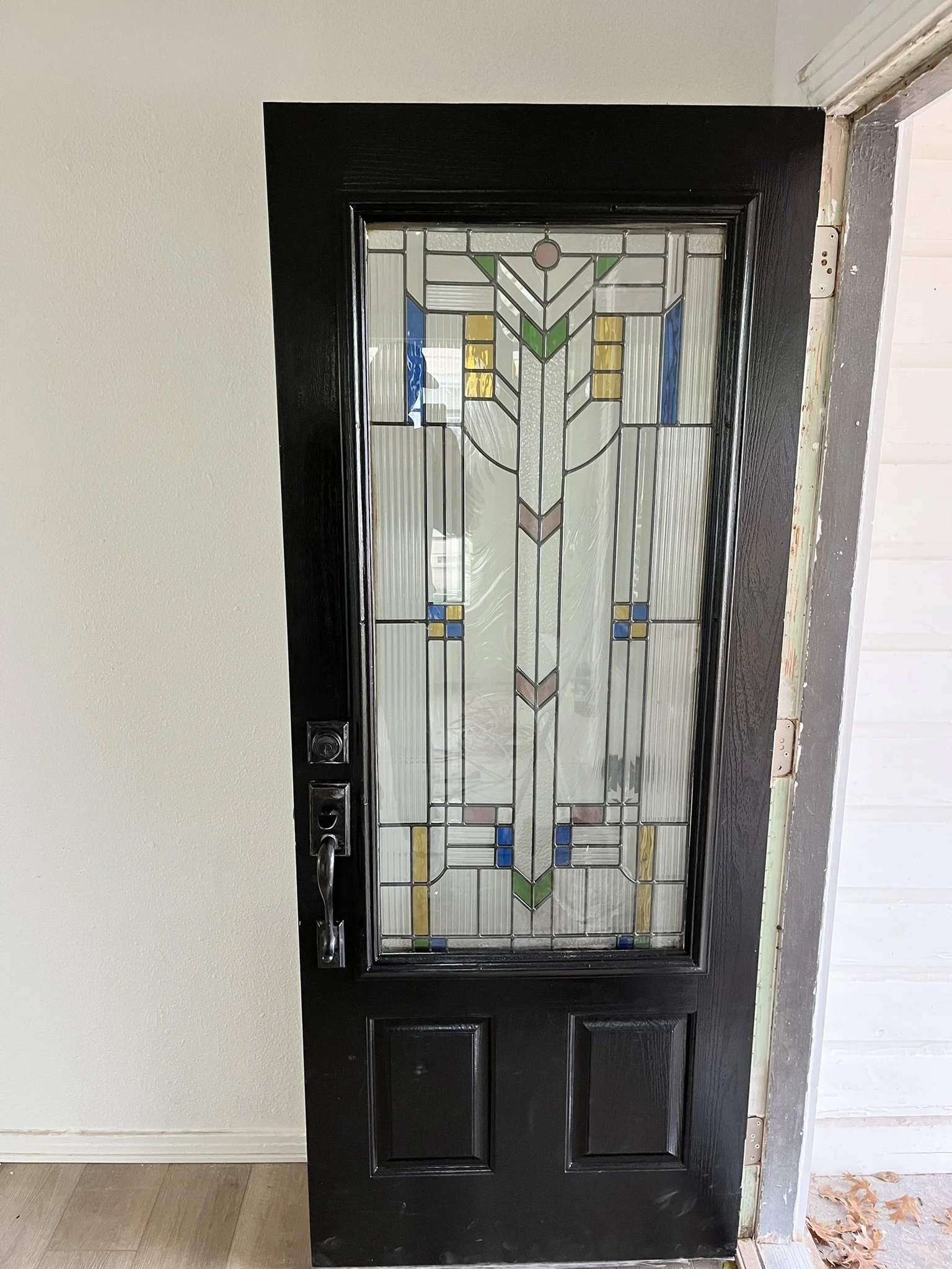A black front door with a stained glass window featuring geometric patterns and colors like green, yellow, blue, and pink, installed in a doorway with a white wall.