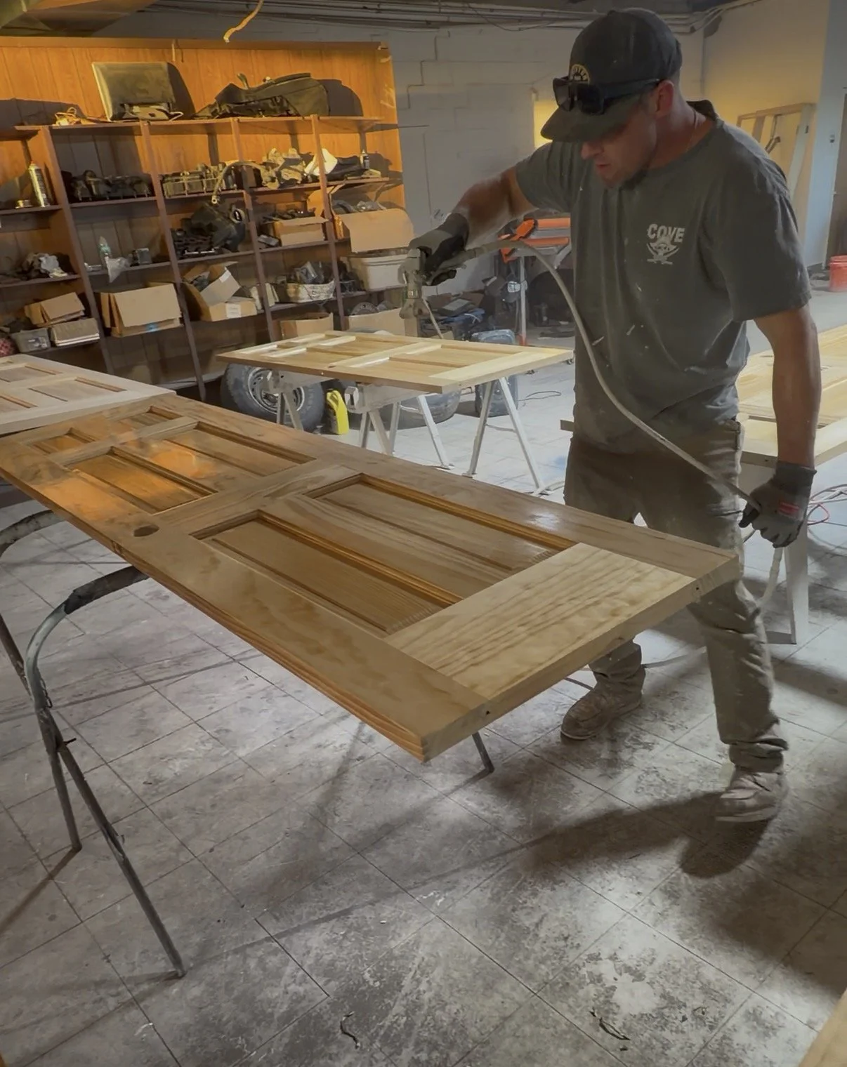 A man wearing a cap, sunglasses, and gloves is spray painting a wooden door in a workshop. The door has four rectangular indentations and is placed on sawhorses. Shelves with tools and supplies are visible in the background.