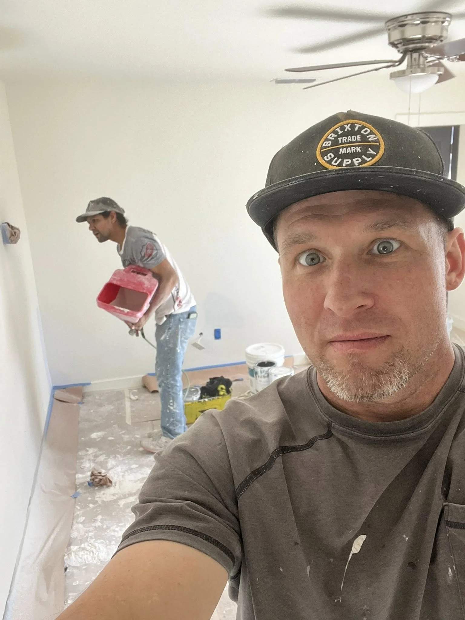 Two men working on painting a room, one in the foreground taking a selfie, wearing a gray shirt and a black cap, the other in the background holding a paint tray, wearing a gray shirt and a gray cap.