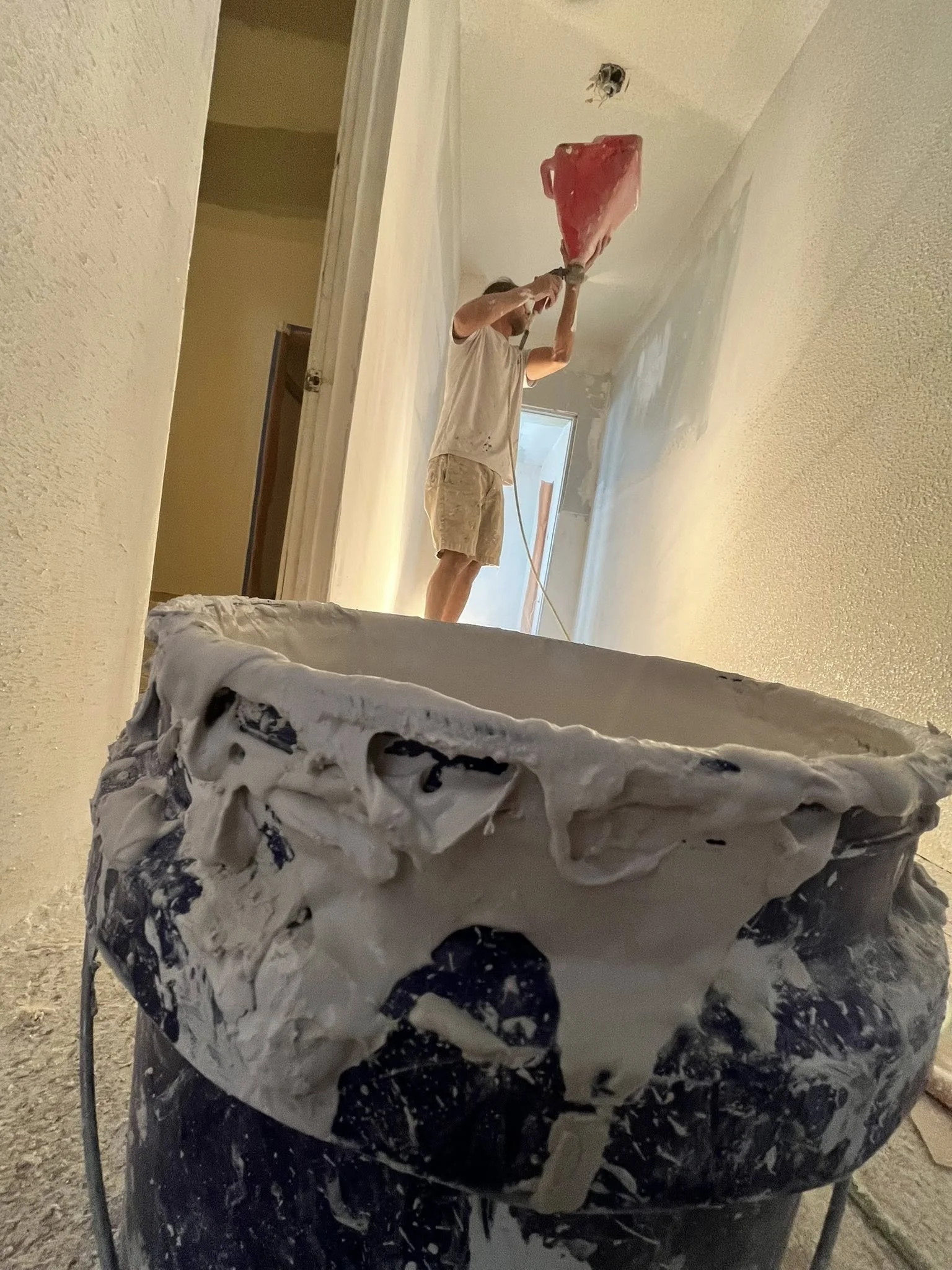 A person painting an interior wall with a roller, standing on a ladder, in a room under renovation, with a bucket of paint in the foreground.