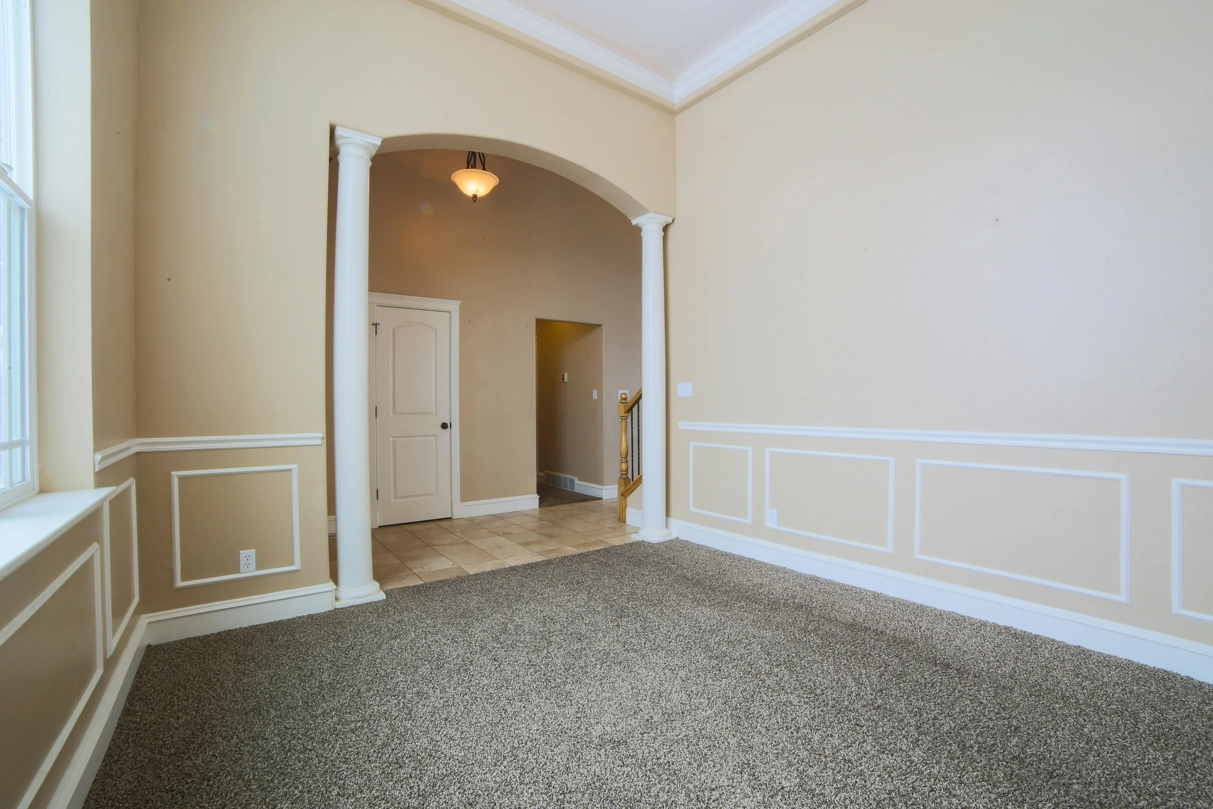 Empty living room with beige walls, carpeted floor, and white trim, leading to an entryway with tile floor and a doorway.
