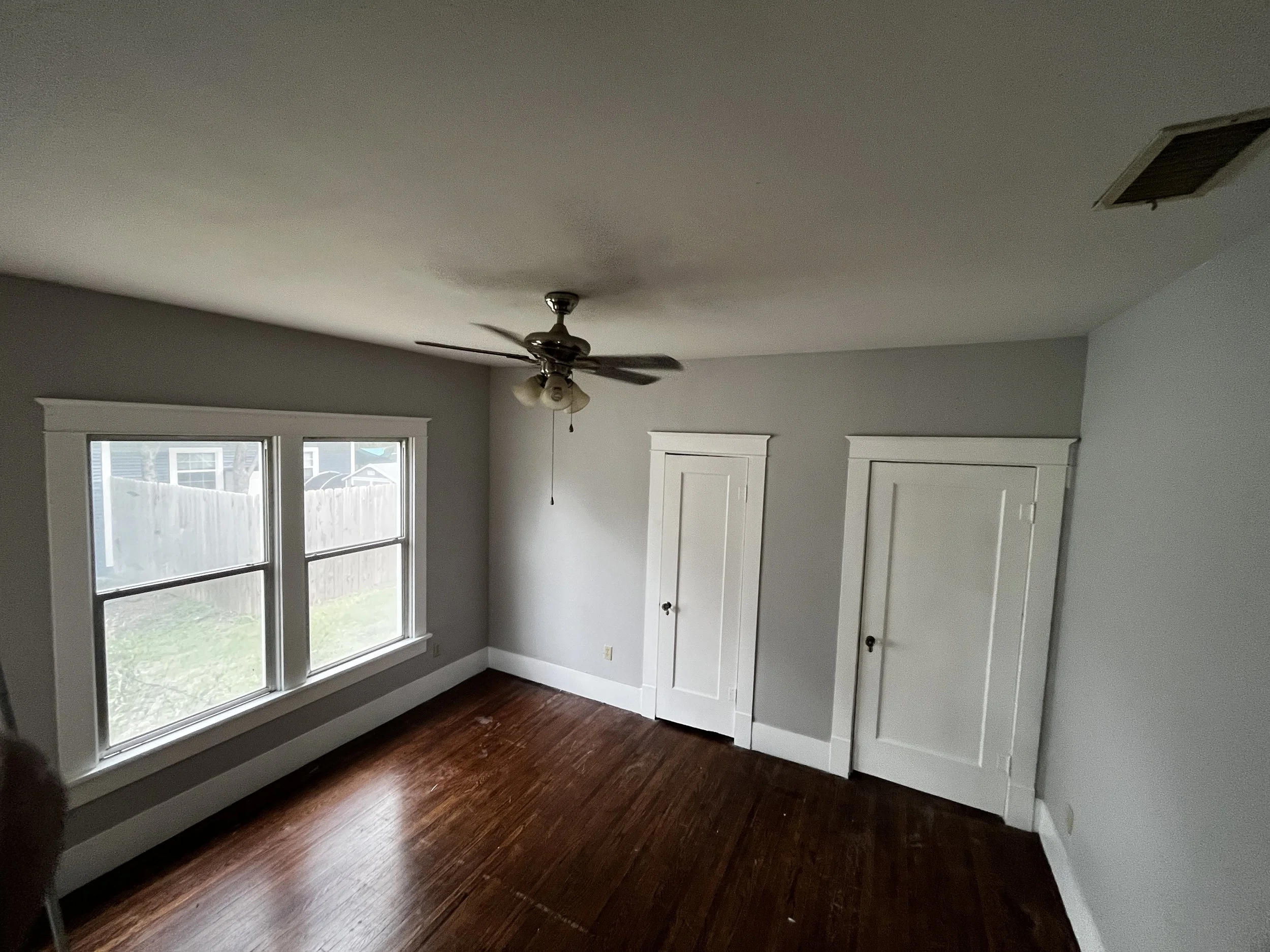 A room with light gray walls, a white ceiling with a ceiling fan and light fixture, three large windows with white trim, and a set of white double closet doors. The floor is dark wood.