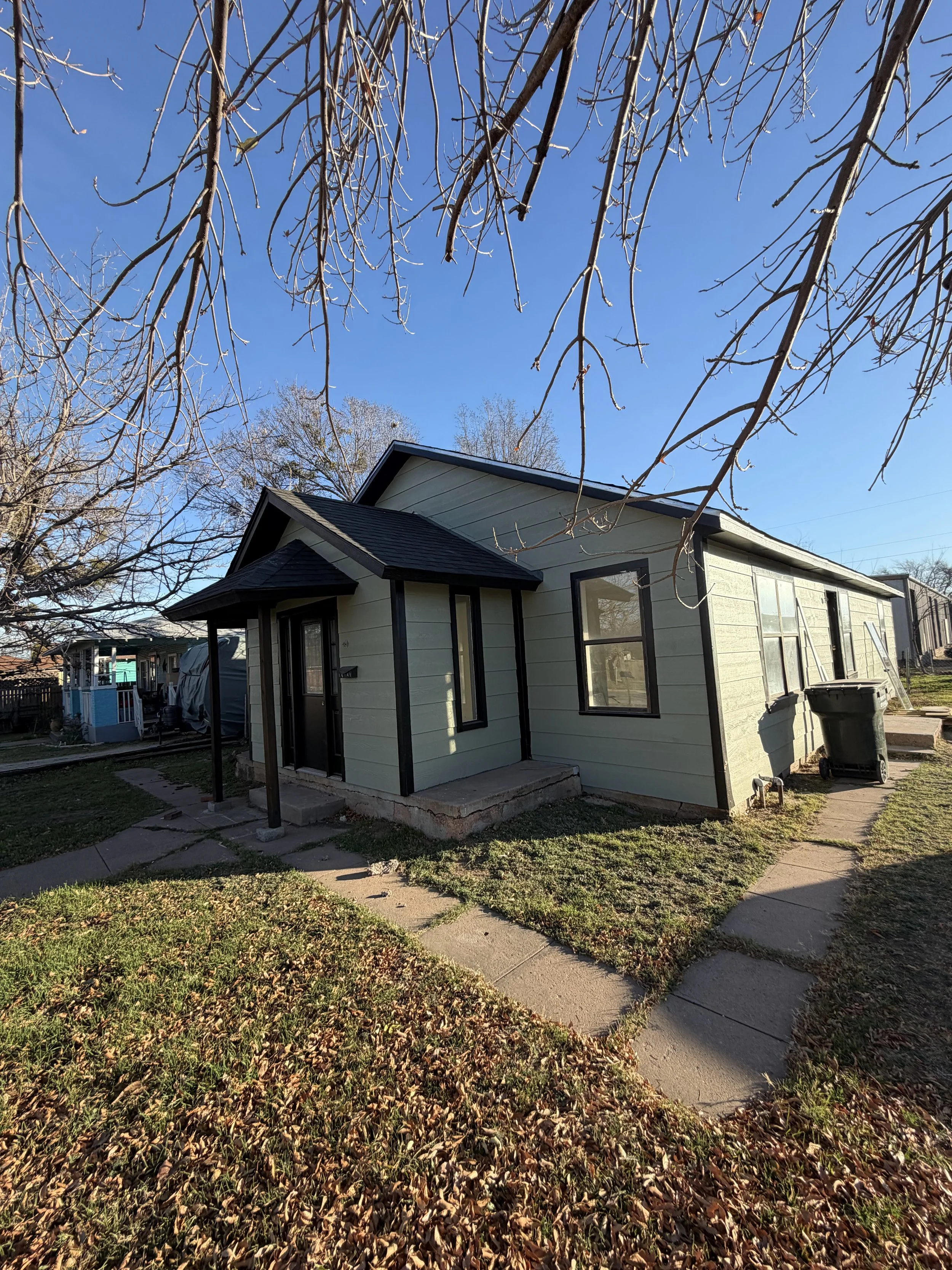 Small house with dark roof, light green siding, and a small front porch, surrounded by a grassy yard with fallen leaves, overhanging tree branches, and a clear blue sky.