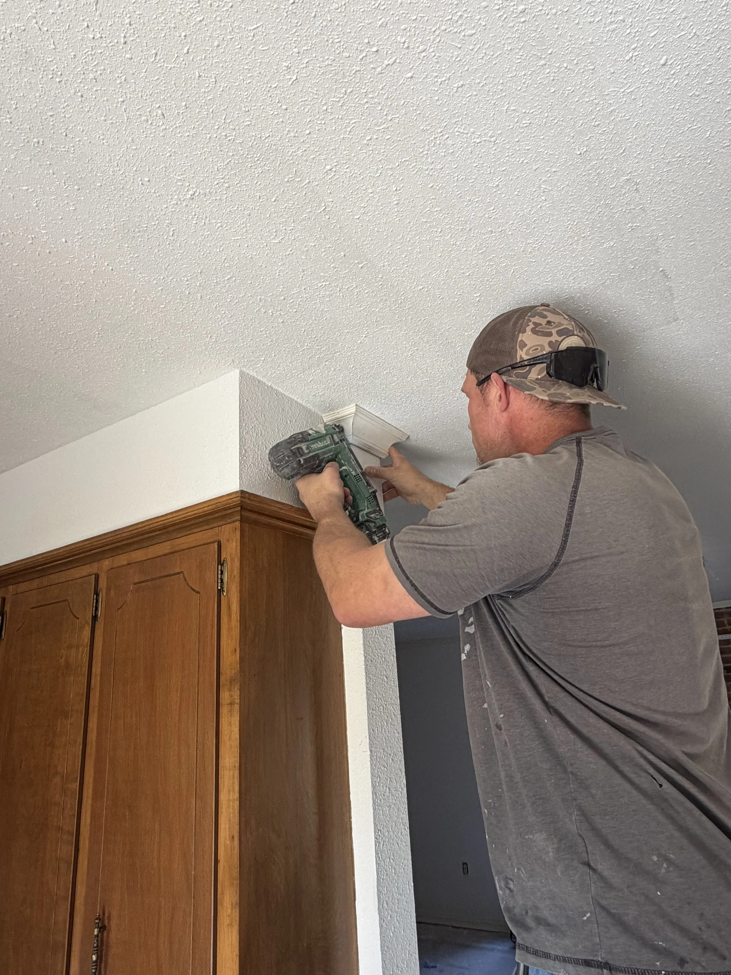 Man installing a decorative crown molding on a ceiling with a cordless drill in a room with white textured ceiling and wooden cabinets.