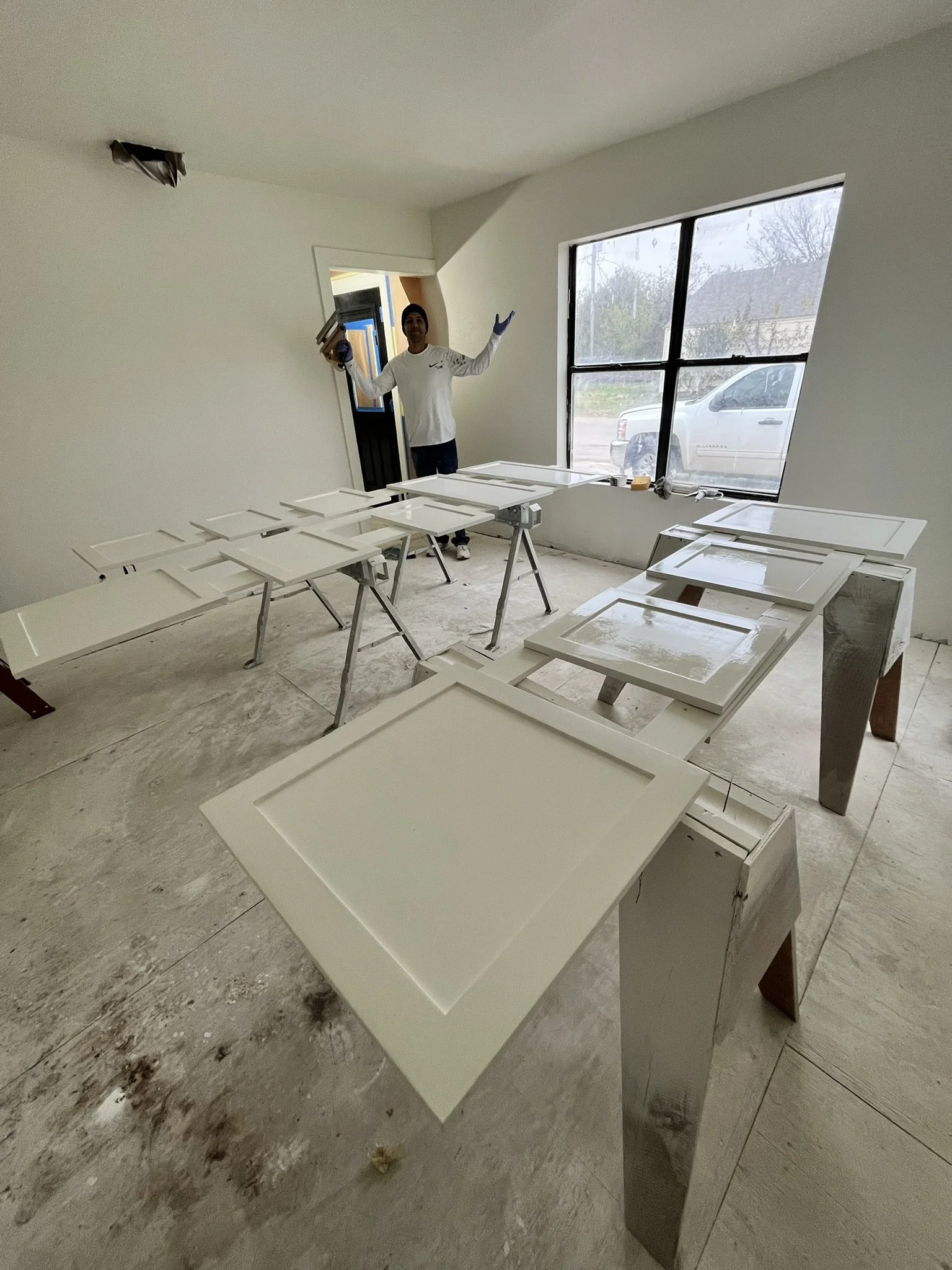 A person standing inside a room with white walls, holding a paint roller and smiling, surrounded by white painted cabinet doors on sawhorses, preparing for installation or painting.