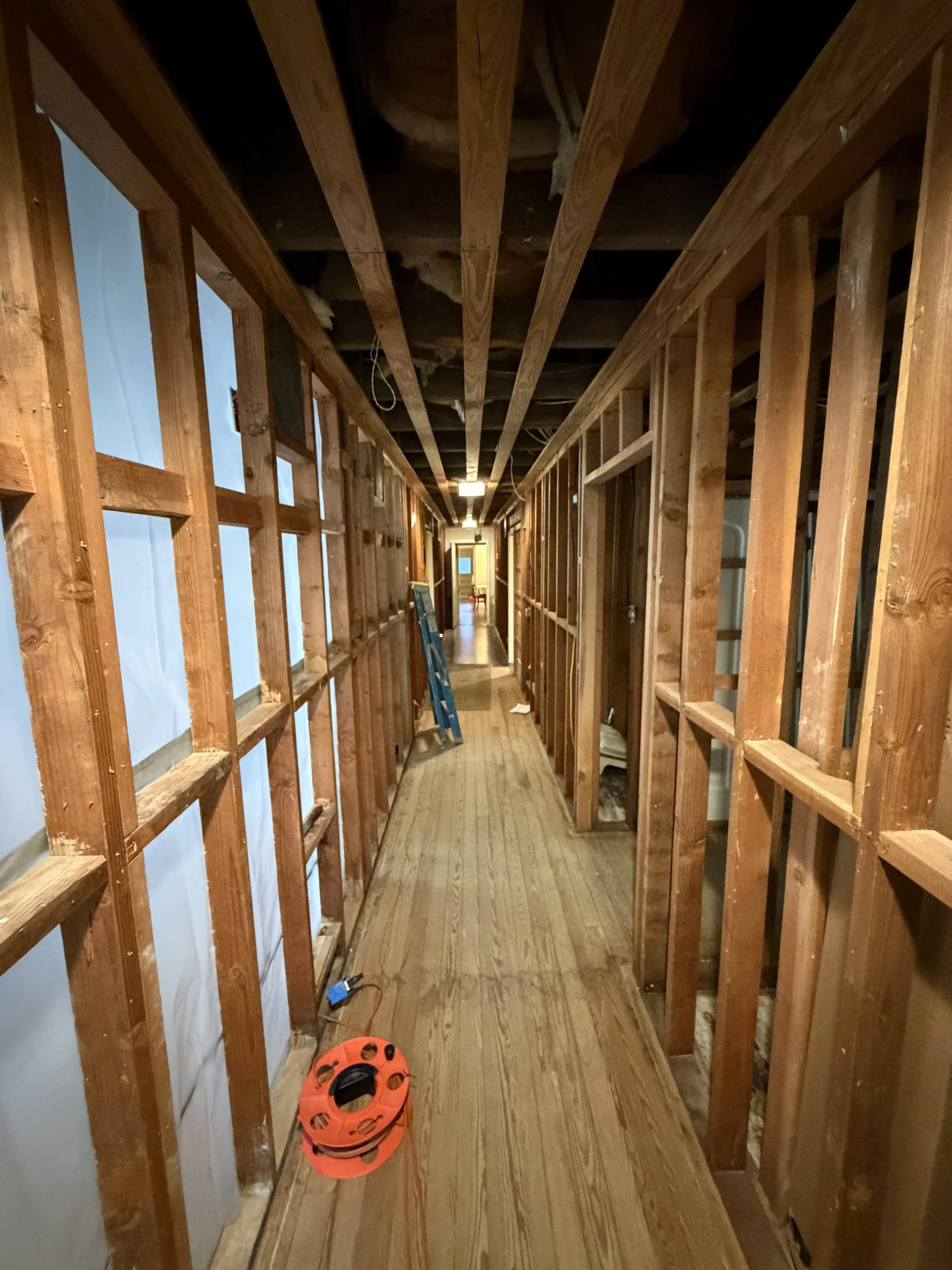 A hallway in a house under renovation with exposed wooden studs and ceiling. Construction tools are on the floor, including a stack of orange helmets. The hallway has wooden flooring and electric wiring hanging from the ceiling.