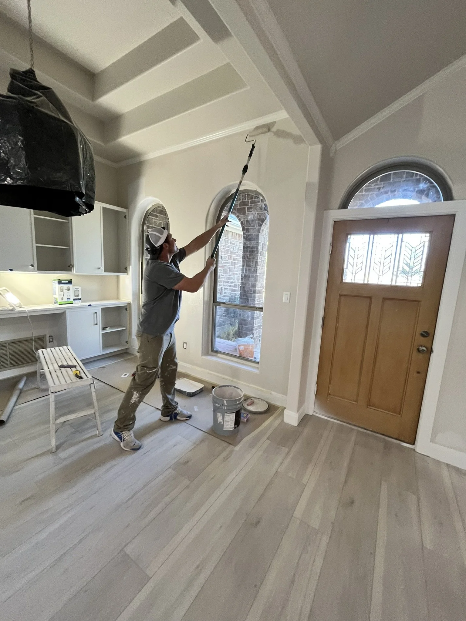 Person painting the ceiling of a room with a roller on an extended pole, standing on the floor near a door and windows in a house interior.