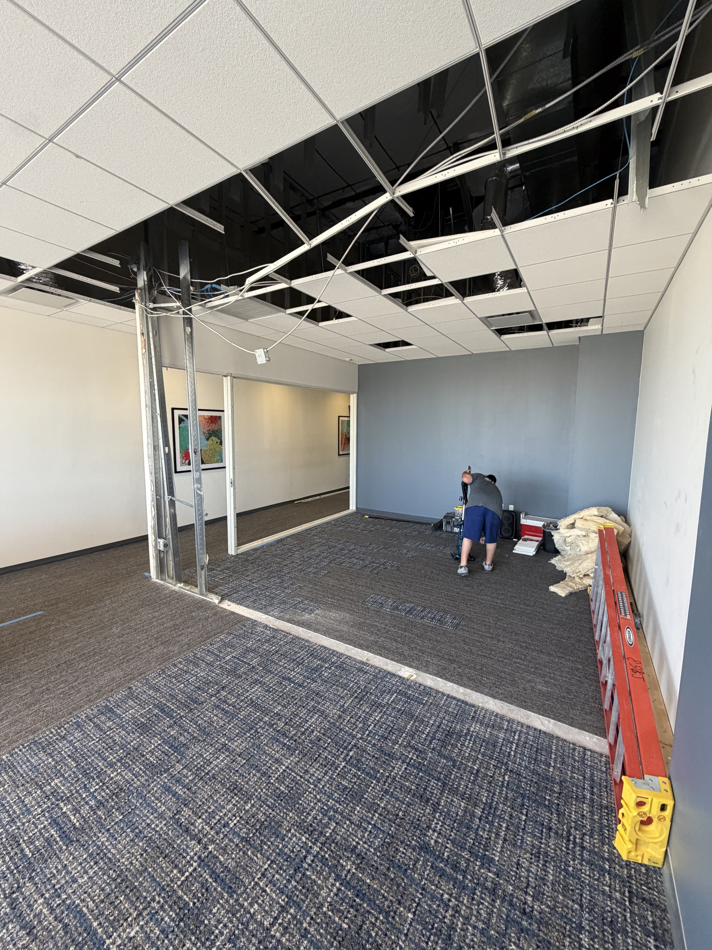 Office ceiling under renovation with missing tiles, visible wiring, and a worker installing or repairing the ceiling panels.