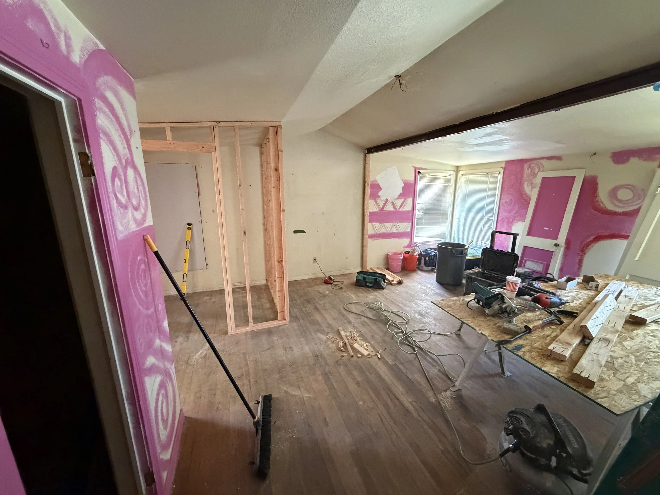 Room under renovation with unfinished walls, construction tools, and materials scattered on the floor, including a wooden frame, buckets, and a worktable.