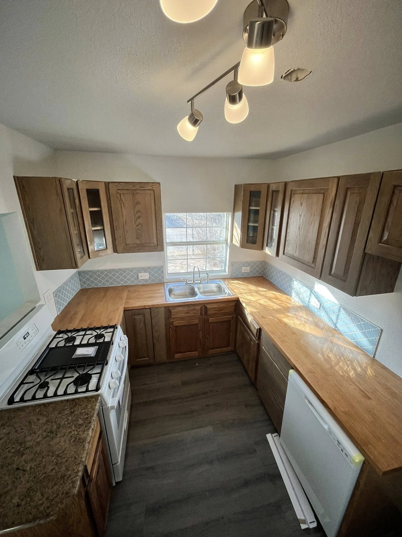 An overhead view of a kitchen with wooden cabinets, a double sink under a window, a white stove, and a white dishwasher. The kitchen features brown countertops, blue patterned backsplash tiles, and dark flooring. A ceiling light fixture with four bulbs illuminates the space.