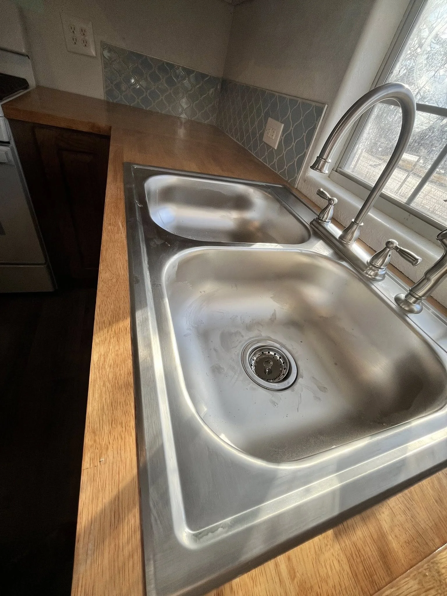 A kitchen countertop with a stainless steel double sink, a window above the sink, a wooden surface, and a backsplash with a patterned tile design.