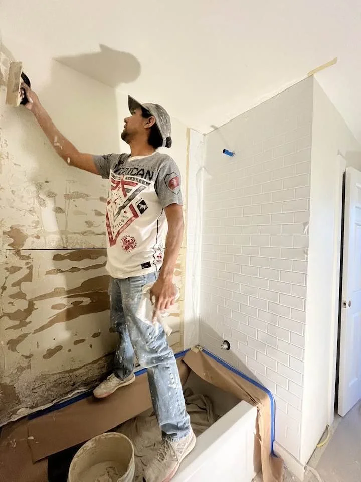 A man standing on a cardboard and plastic covered bathtub, painting a wall with a drywall compound or joint compound, in a room under renovation.