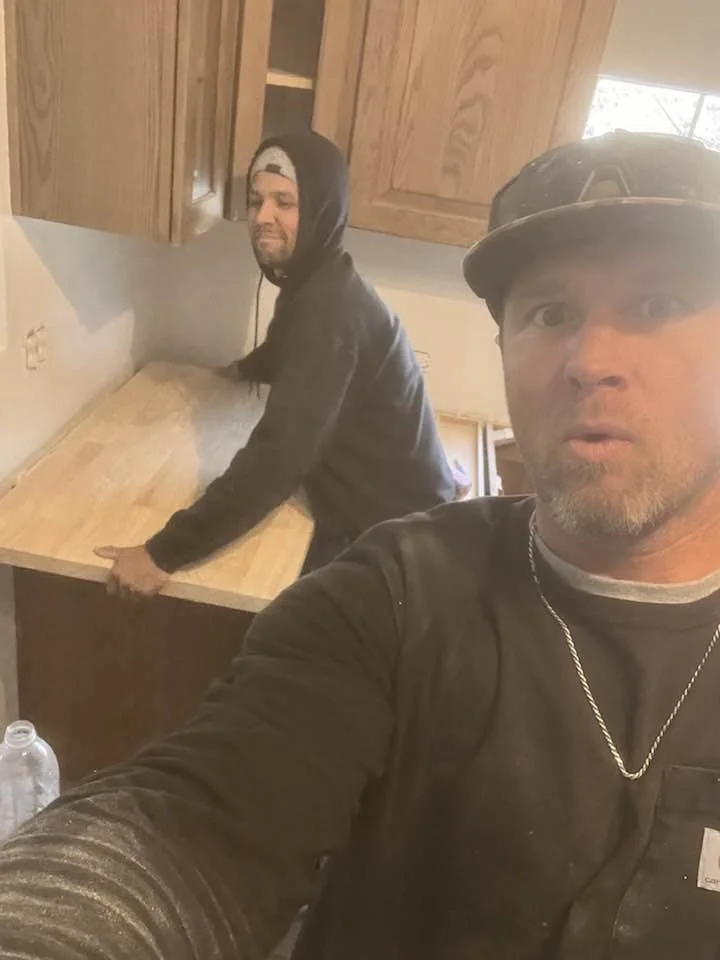 Two men in black hoodies in a kitchen with wooden cabinets. One man is in the foreground wearing a cap and taking a selfie, and the other man is in the background leaning over a wooden countertop.