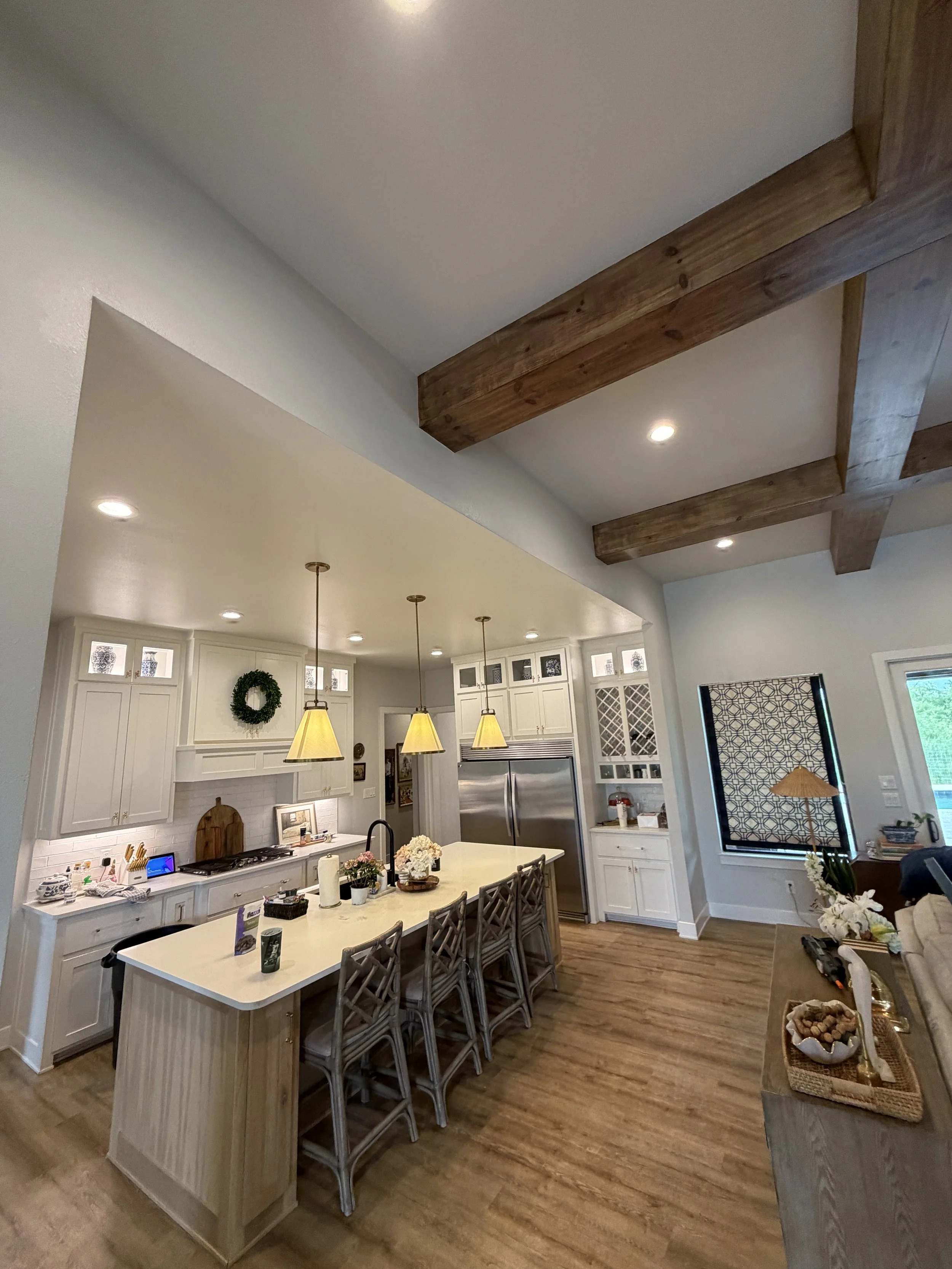 A cozy kitchen with white cabinets, island counter, wooden beams on the ceiling, and a dining table with chairs. Pendant lights hang above the island, and there is a window with a black patterned shade. Decorations include a wreath on the wall and fl