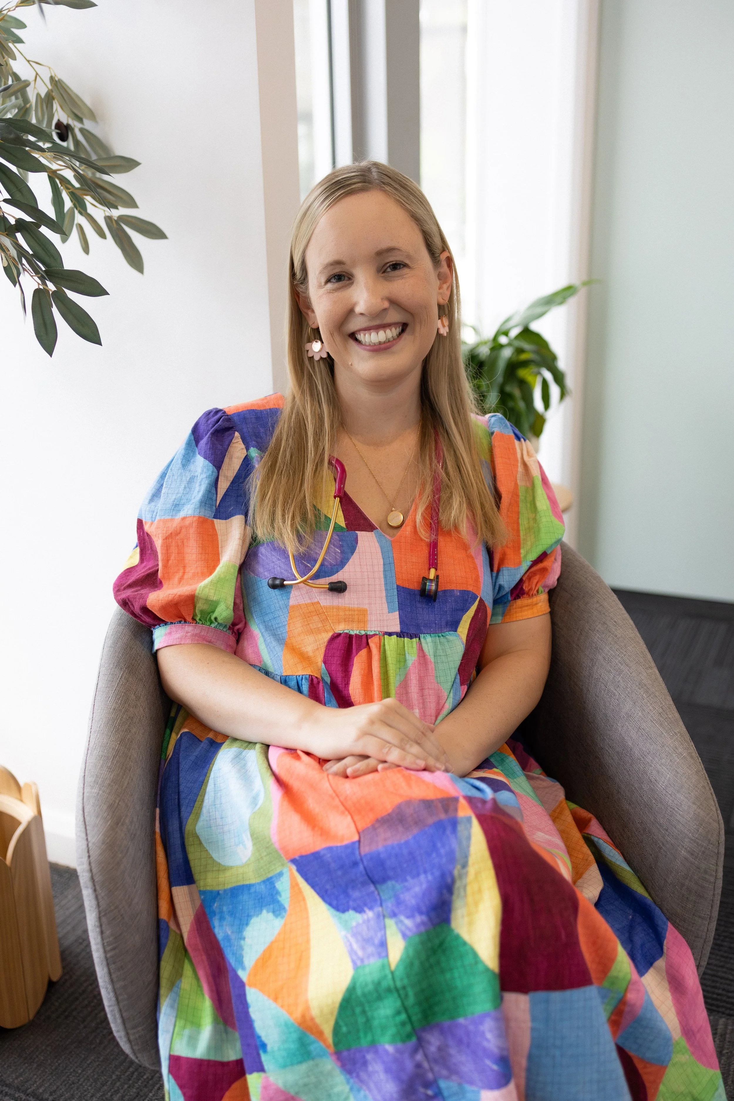 A smiling woman with blonde hair seated in a fabric armchair, wearing a multicolored patchwork dress, pink earrings resembling flowers, and a necklace, in a bright room with plants and windows.