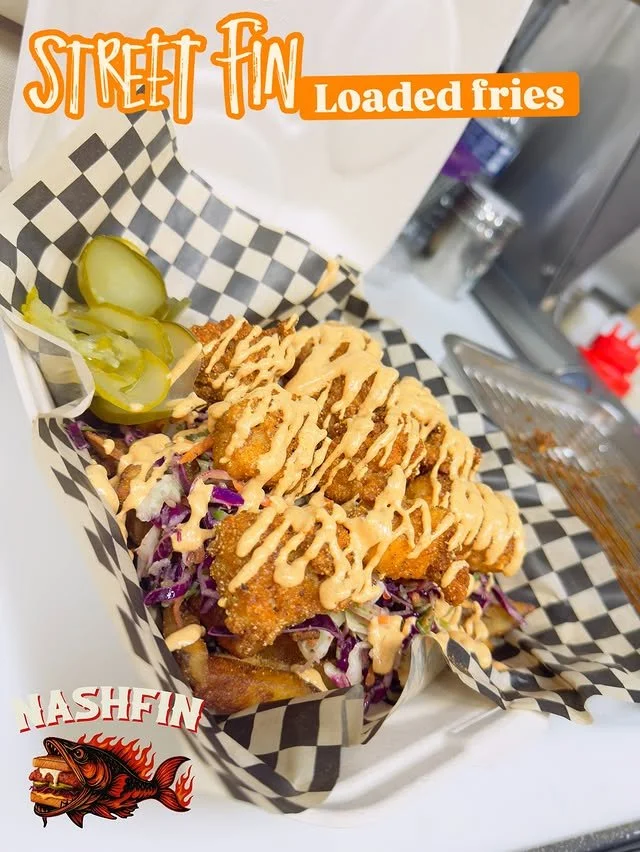 Loaded fried fish tacos topped with a creamy sauce, served on a checkered paper in a white basket, with pickles on the side.