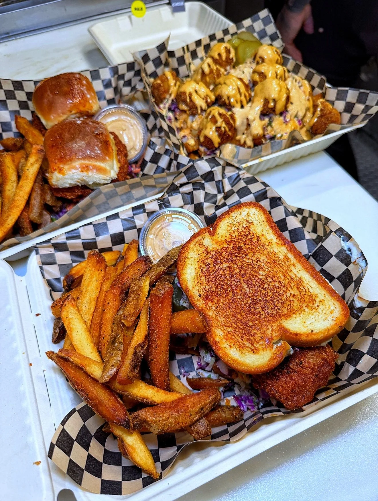 Two takeout containers with fries, fried chicken sandwiches with toasted buns, and a basket of fried food topped with sauce.