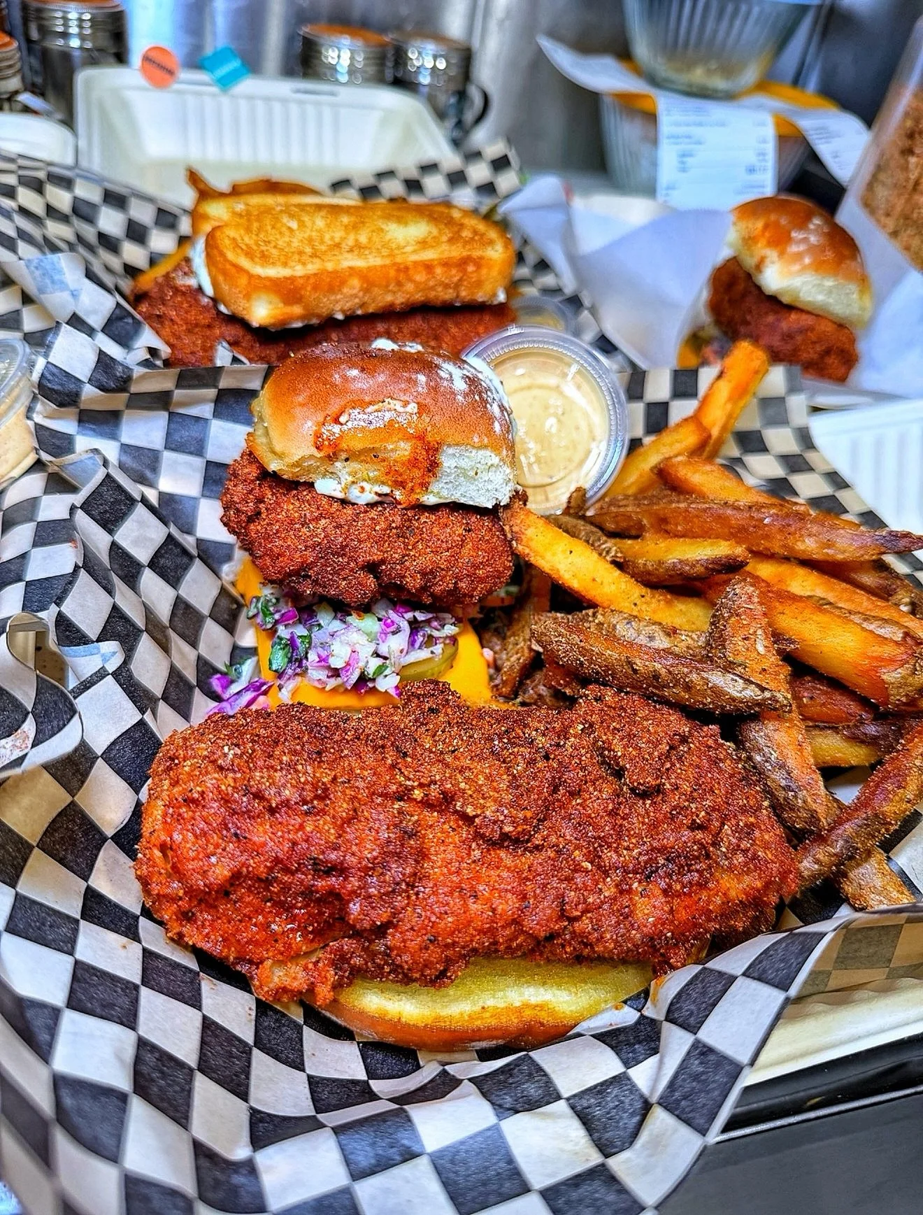 Fried chicken sandwich with coleslaw and fries, served with a side of dipping sauce, on a checkered paper-lined tray.