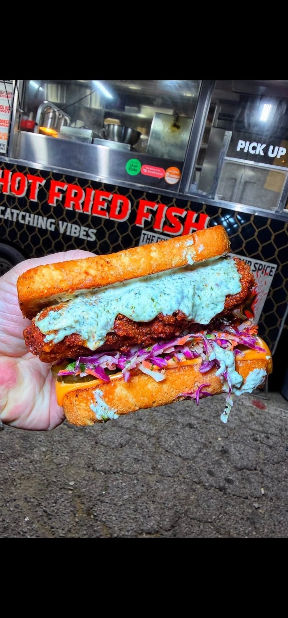 Close-up of a hand holding a fried fish sandwich with lettuce, tomato, coleslaw, tartar sauce, and a toasted hoagie bun, with a food truck in the background that has a sign reading "Hot Fried Fish".