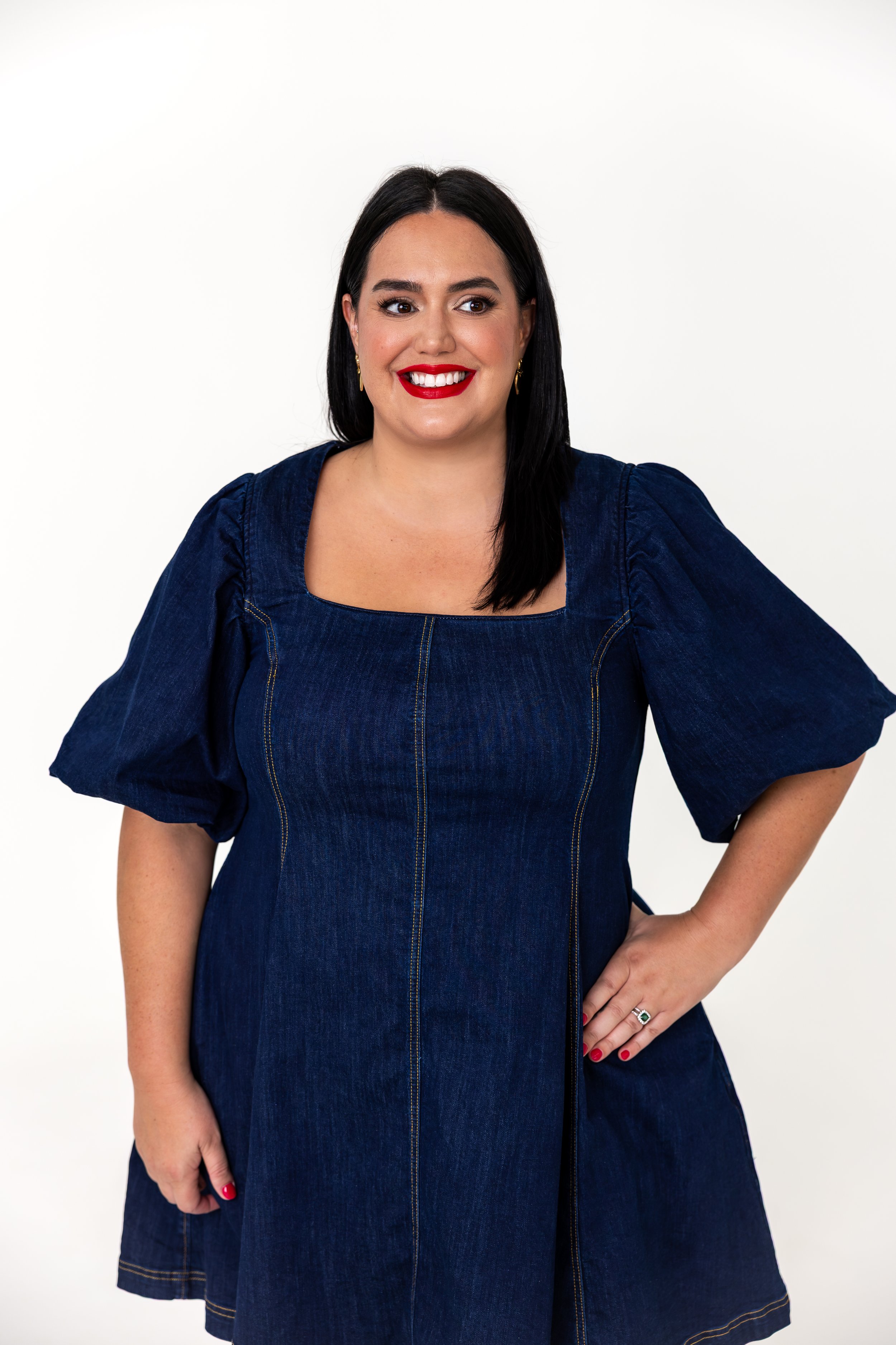 A woman with black hair, red lipstick, and a smile wearing a dark blue denim dress with puffed sleeves, standing against a white background.