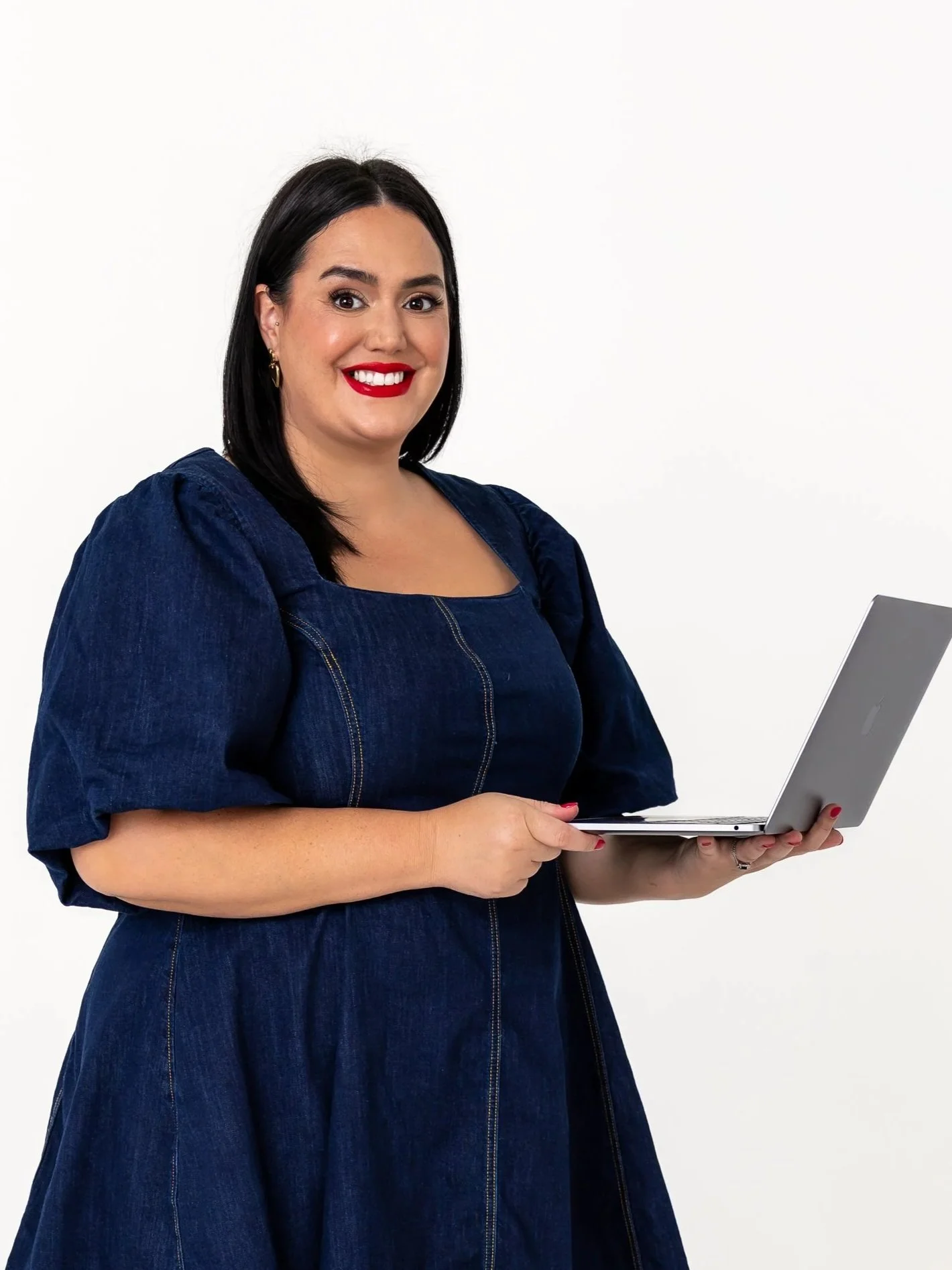 A woman with dark hair, red lipstick, and gold earrings holding an open silver laptop, wearing a dark blue denim dress, standing against a white background.