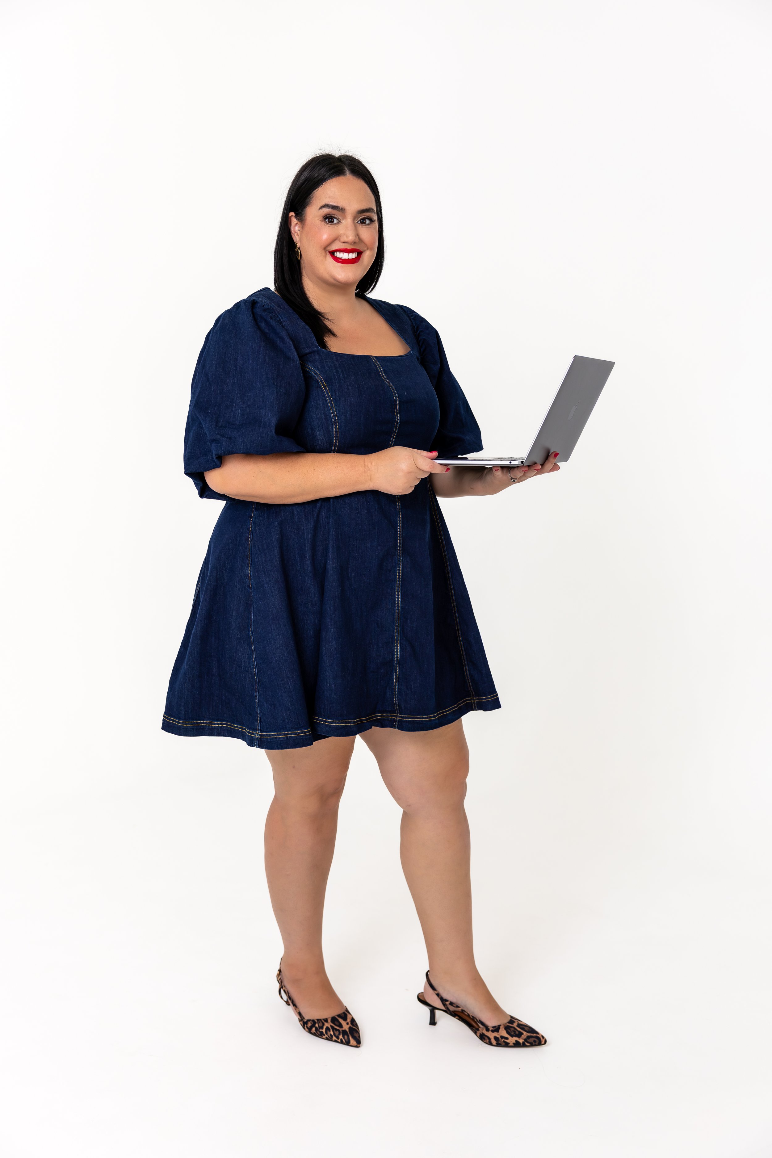 Woman with dark hair, red lipstick, wearing a denim dress with puffed sleeves, leopard print high heels, holding a silver laptop, standing against a plain white background.