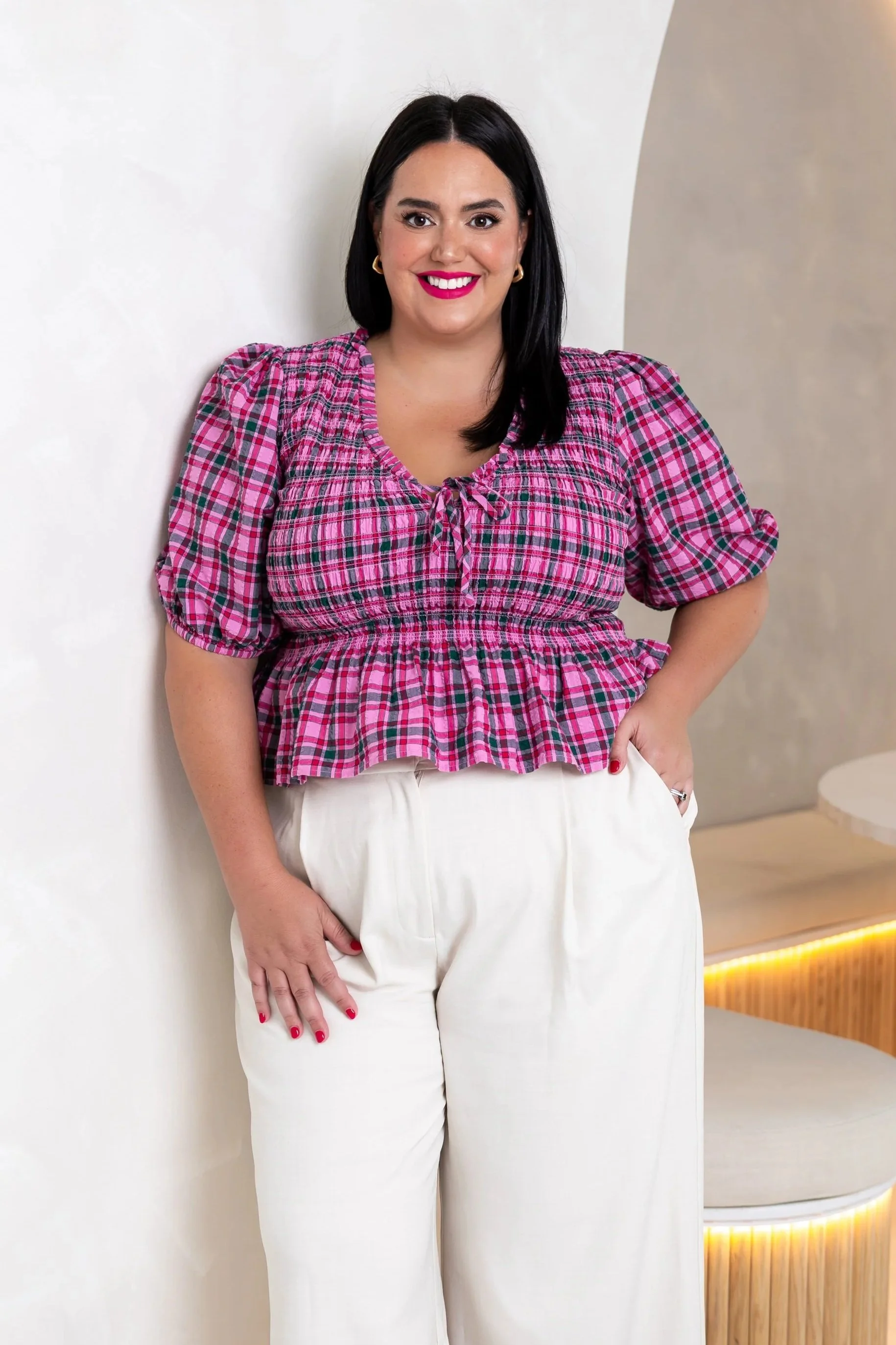 A woman with dark hair, wearing a pink plaid top and white pants, stands against a light-colored wall, smiling with red lipstick.