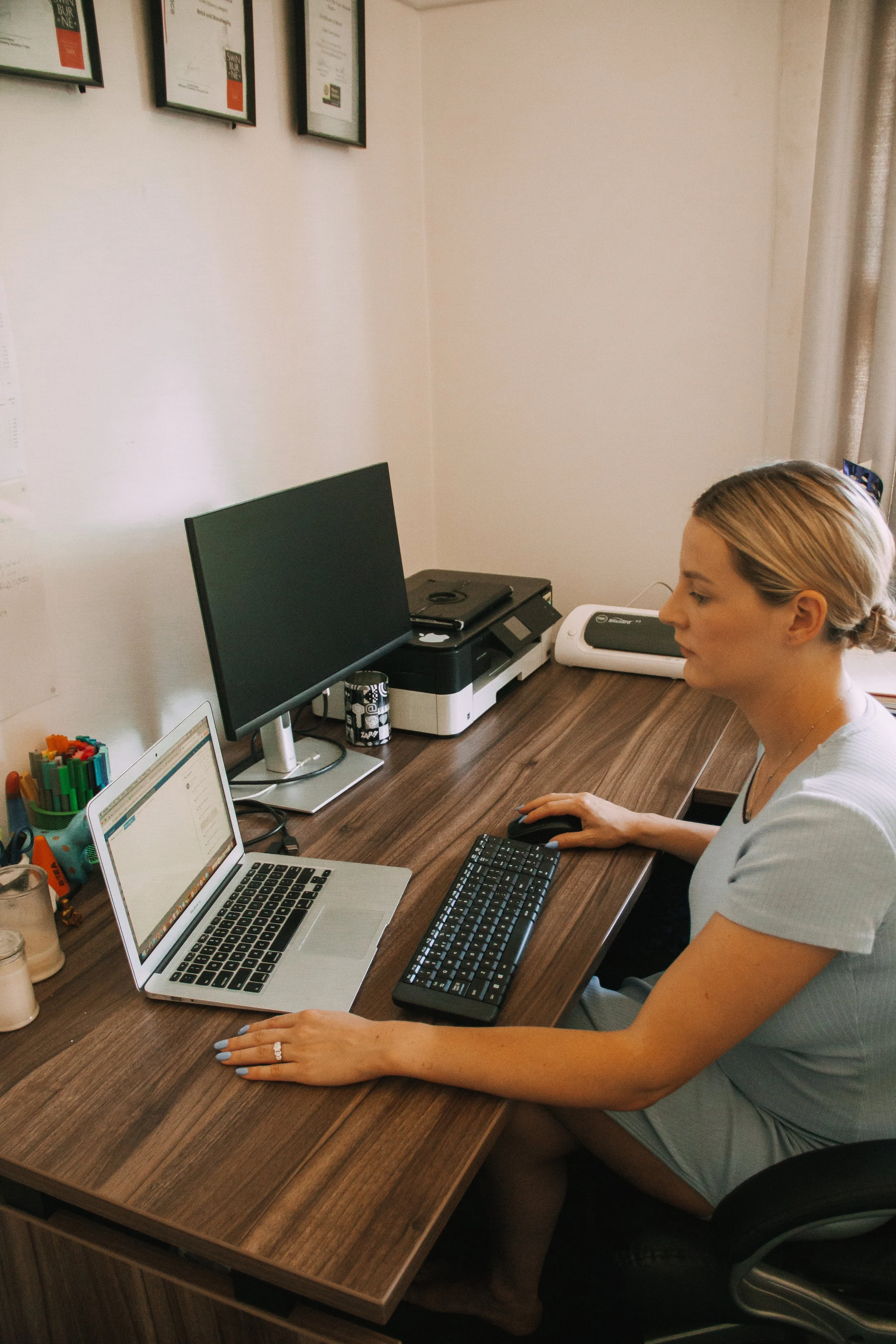 A woman with blonde hair sitting at a wooden desk, working on a computer. The desk has a laptop, a desktop monitor, a keyboard, and a mouse. There are framed certificates on the wall and various office supplies on the desk.