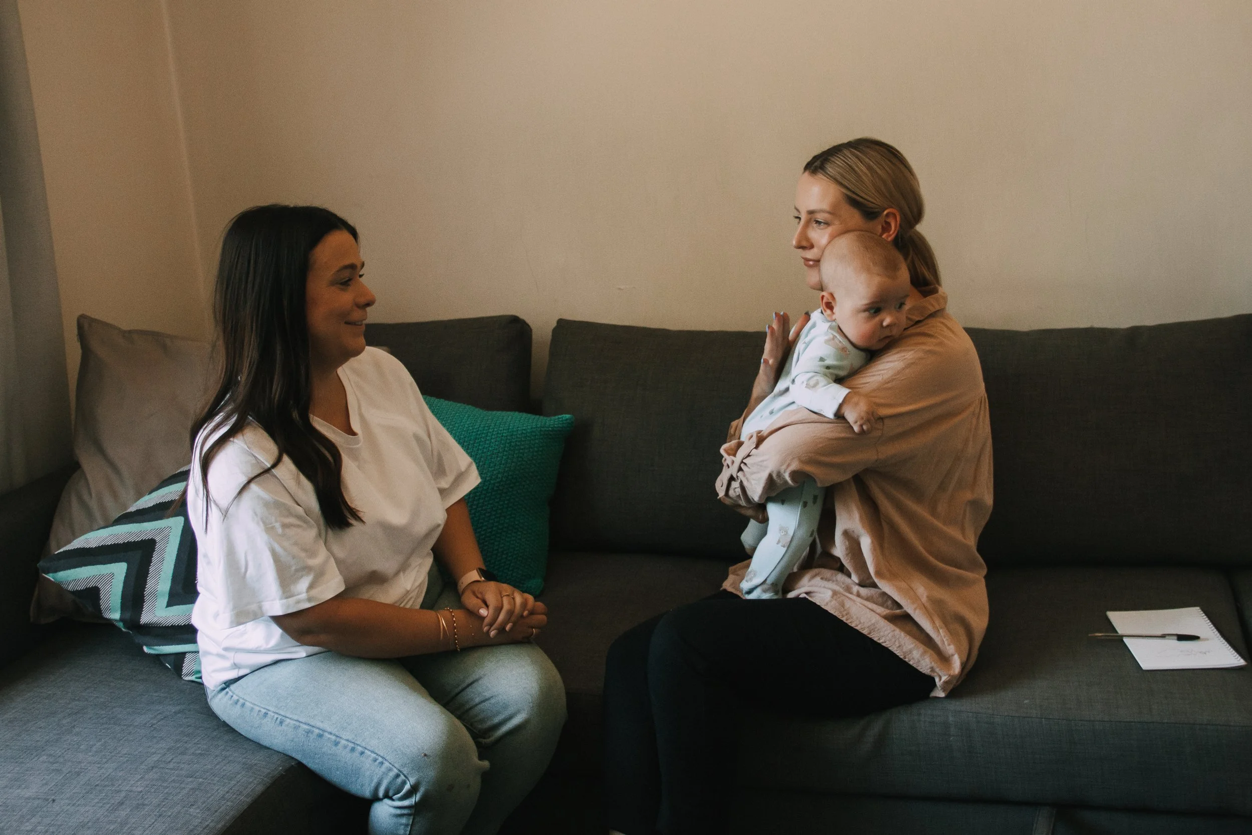 Two women sitting on a couch with a baby. The woman on the left has long dark hair, is wearing a white shirt and light blue jeans, and is smiling at the baby. The woman on the right has blonde hair, is holding the baby, and is smiling at the woman on