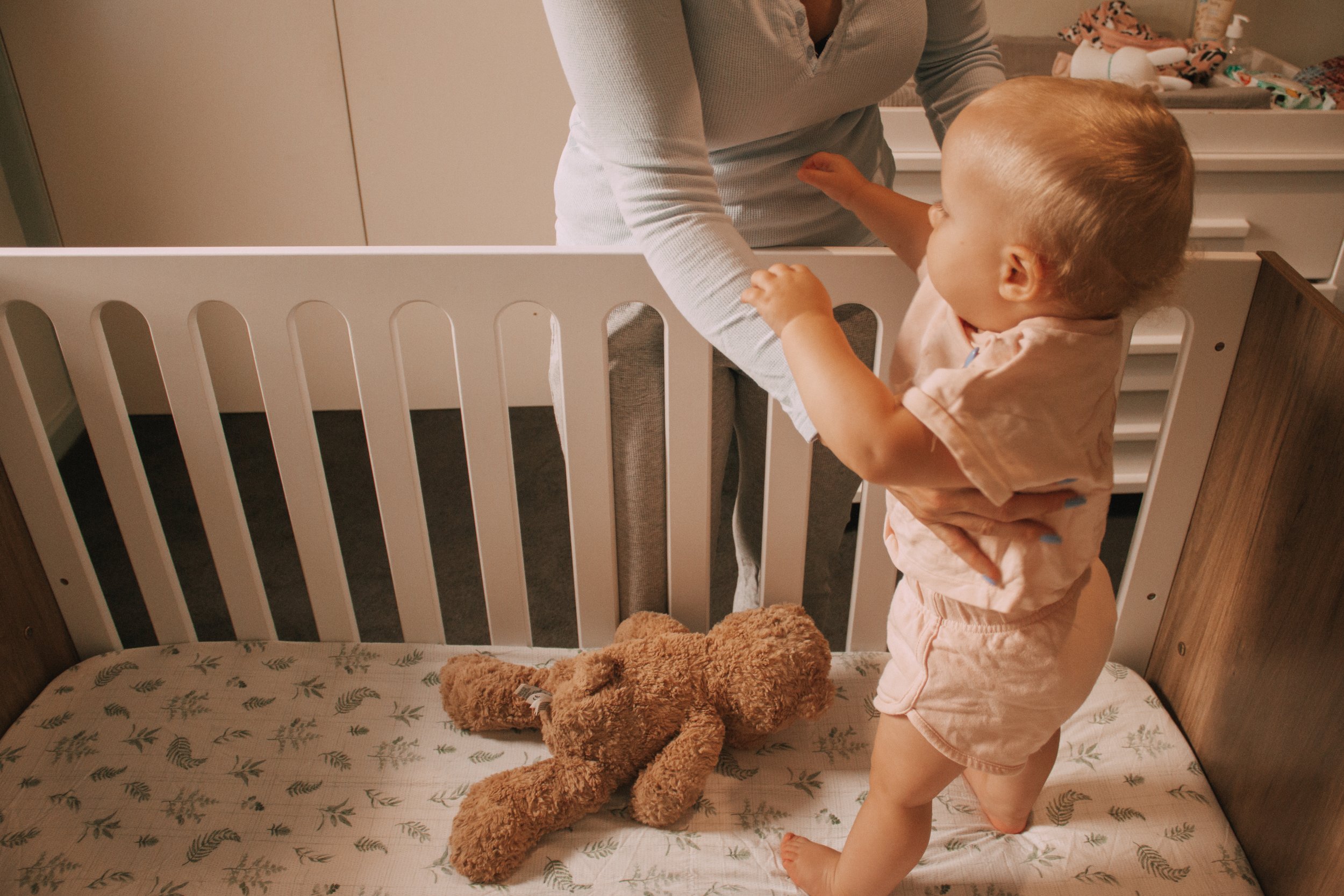 A young child with short hair, wearing light pink pajamas, is standing inside a crib and reaching out to an adult who is leaning over from the outside. The crib has a fitted sheet with a leafy pattern, and a brown plush teddy bear is lying on the mattress. In the background, there are toys and baby supplies on a changing table.