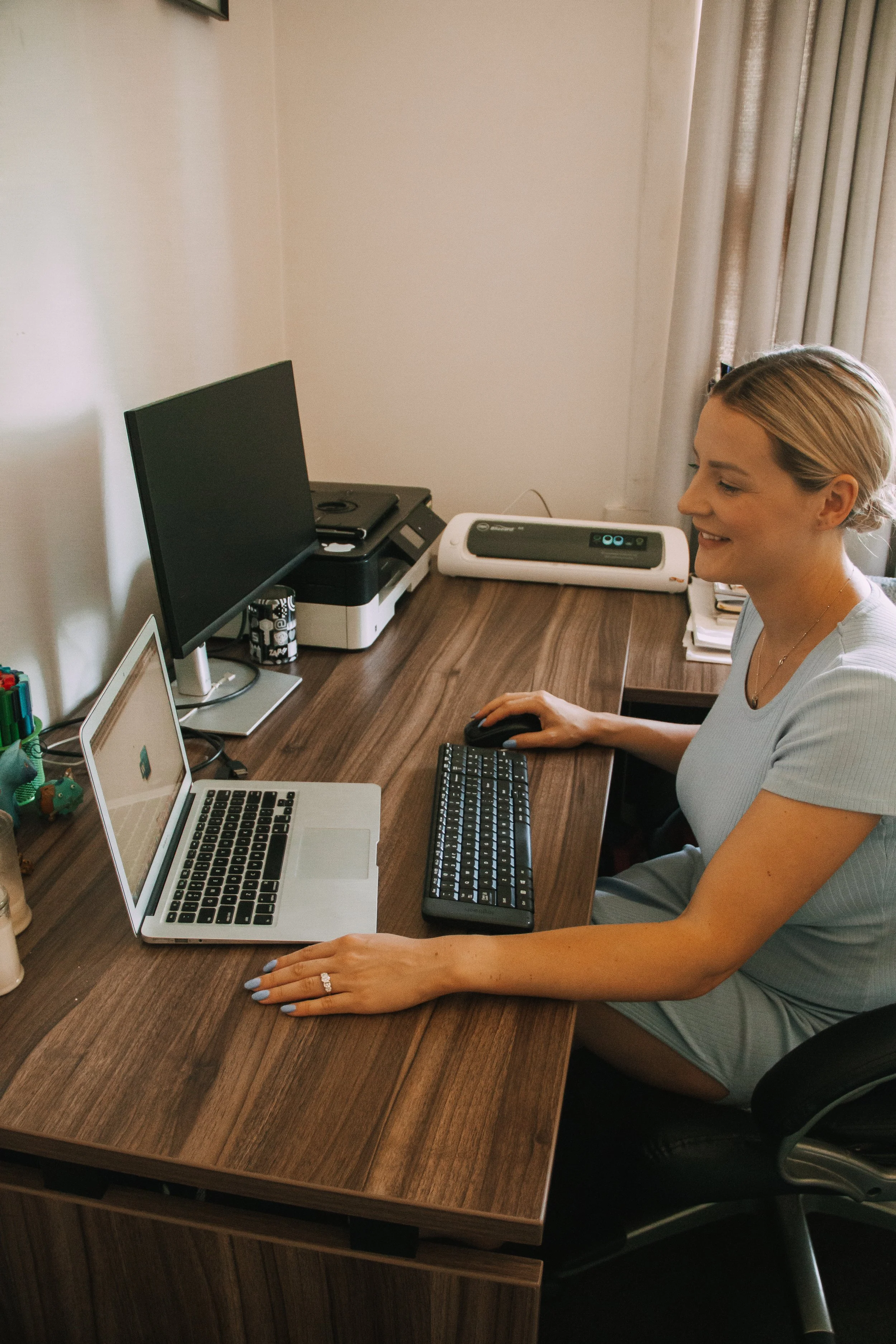 A woman with blonde hair working at a desk, using a computer mouse, with a laptop, keyboard, and printer on the desk, in a room with beige walls and curtains.