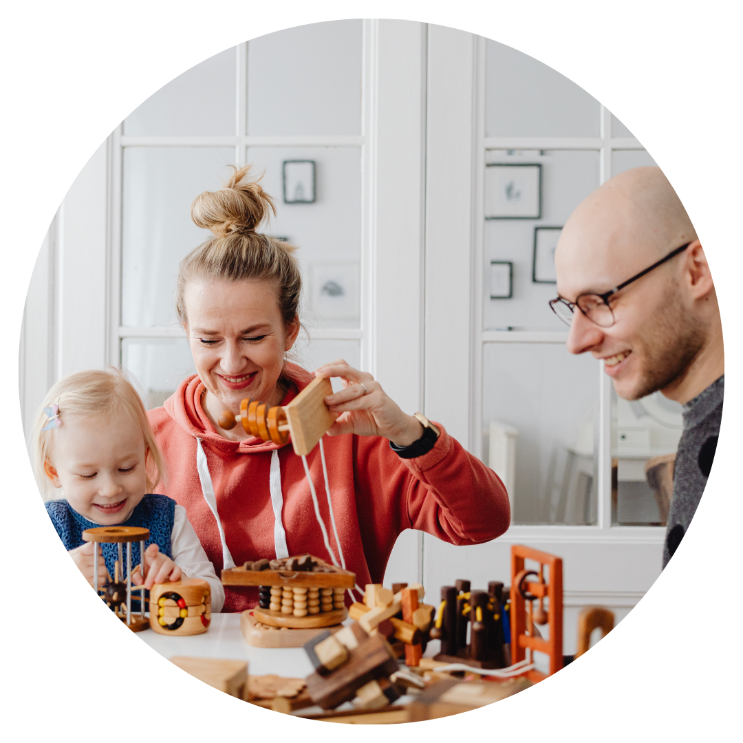 A family of three, including a woman, a young girl, and a man, playing with wooden toys at a table indoors.