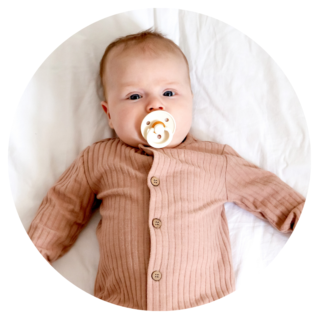 A baby with light skin and light-colored hair lying on a white surface, wearing a brown ribbed shirt with buttons and a pacifier in their mouth.