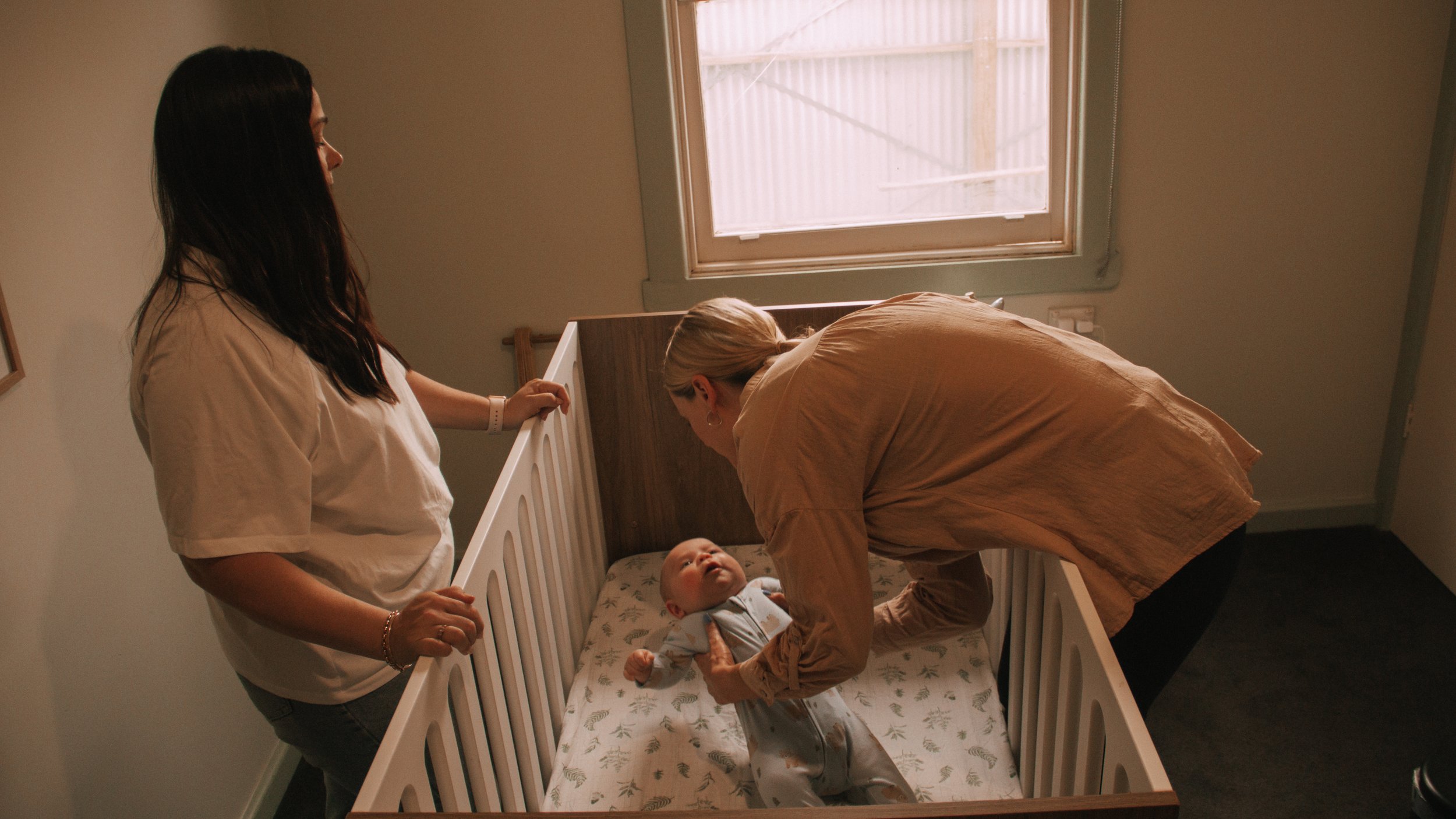 A young baby lying in a crib looking up at two women leaning over, one of whom appears to be the mother, in a nursery room with a window in the background.