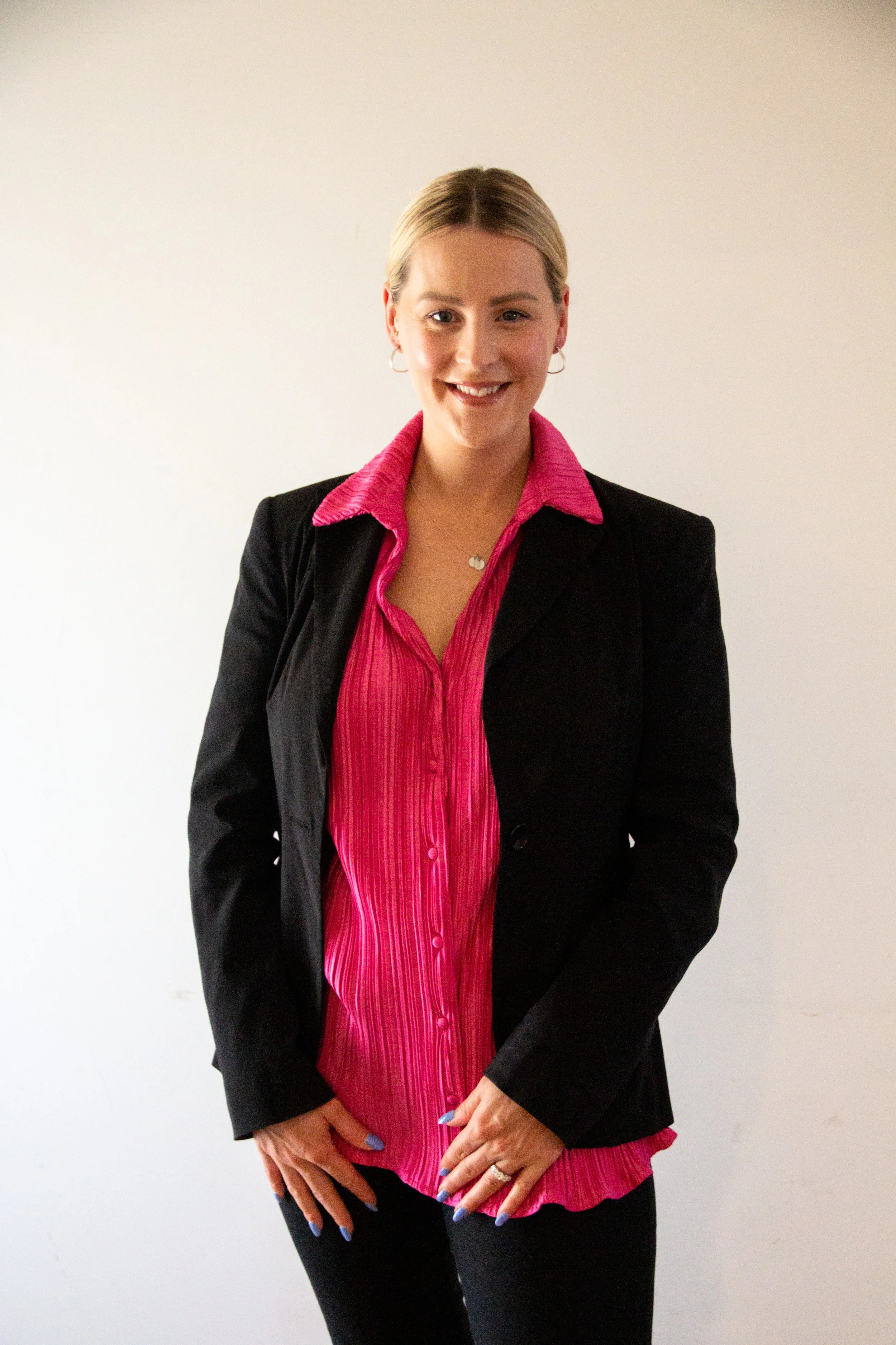 A woman with blonde hair styled in a bun, wearing a black blazer over a pink striped blouse, and black pants, standing against a plain white wall.