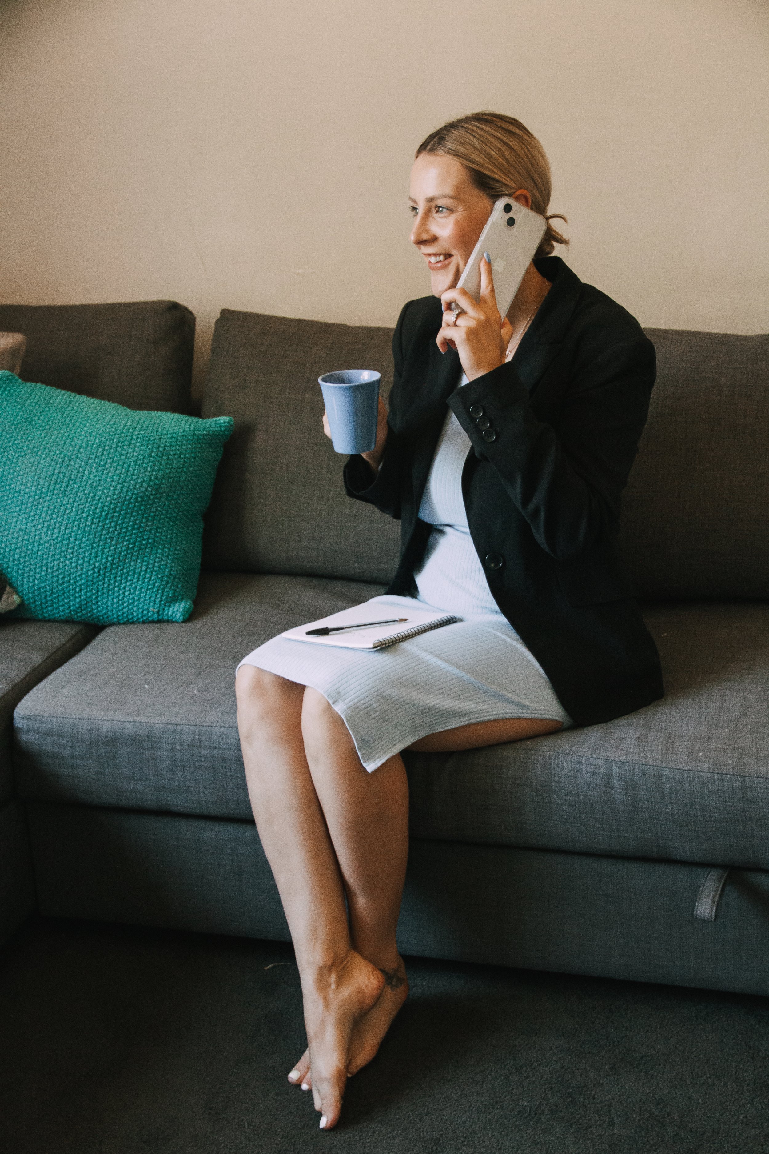 A woman sitting on a gray sofa, holding a phone to her ear with a smile, holding a blue mug in her other hand, with a notebook and pen on her lap, wearing a black blazer and white dress, with her legs crossed, and standing barefoot.