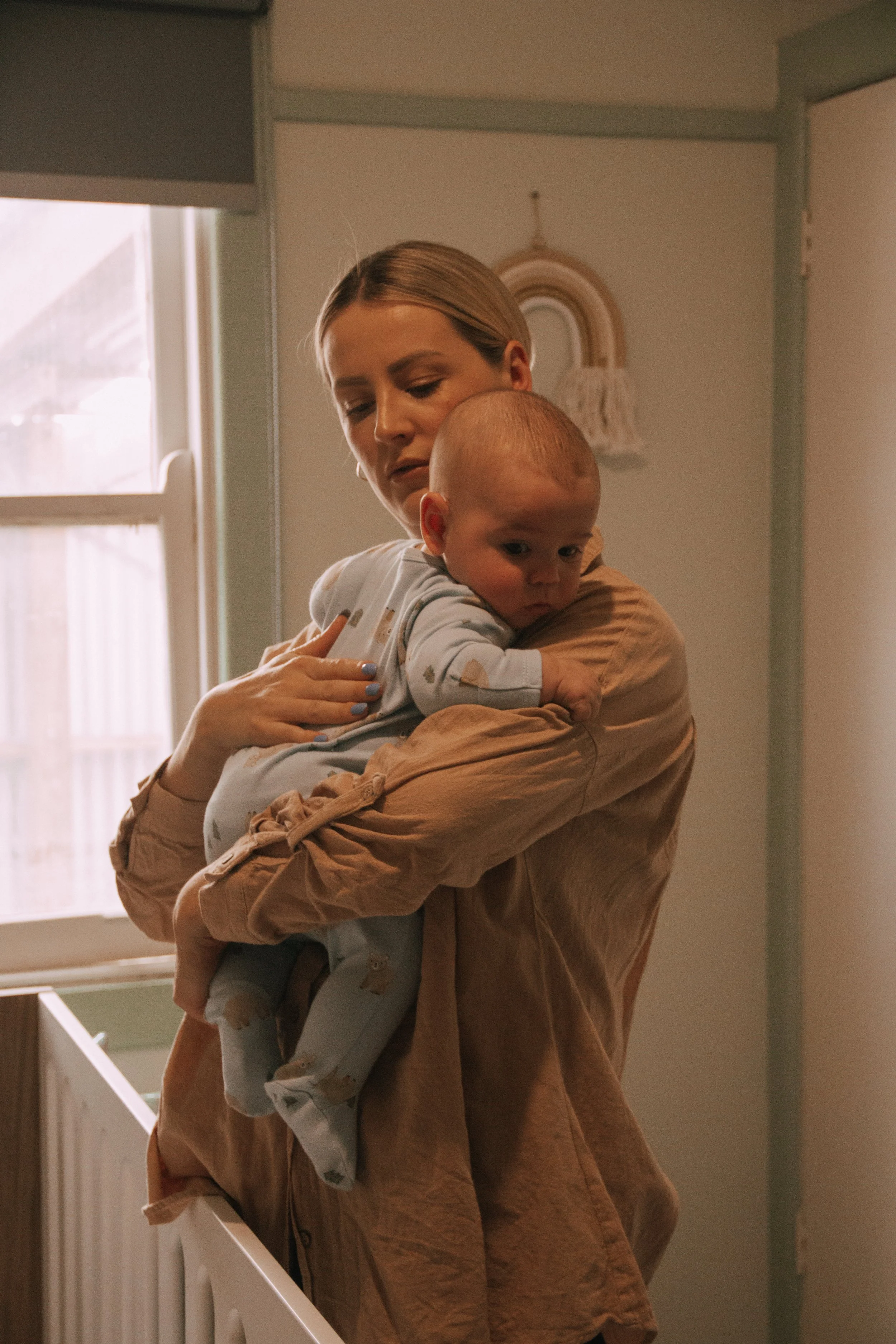 A woman with short blonde hair hugging a baby with a shaved head in a room with natural light coming through a window. There is a rainbow wall hanging behind them.