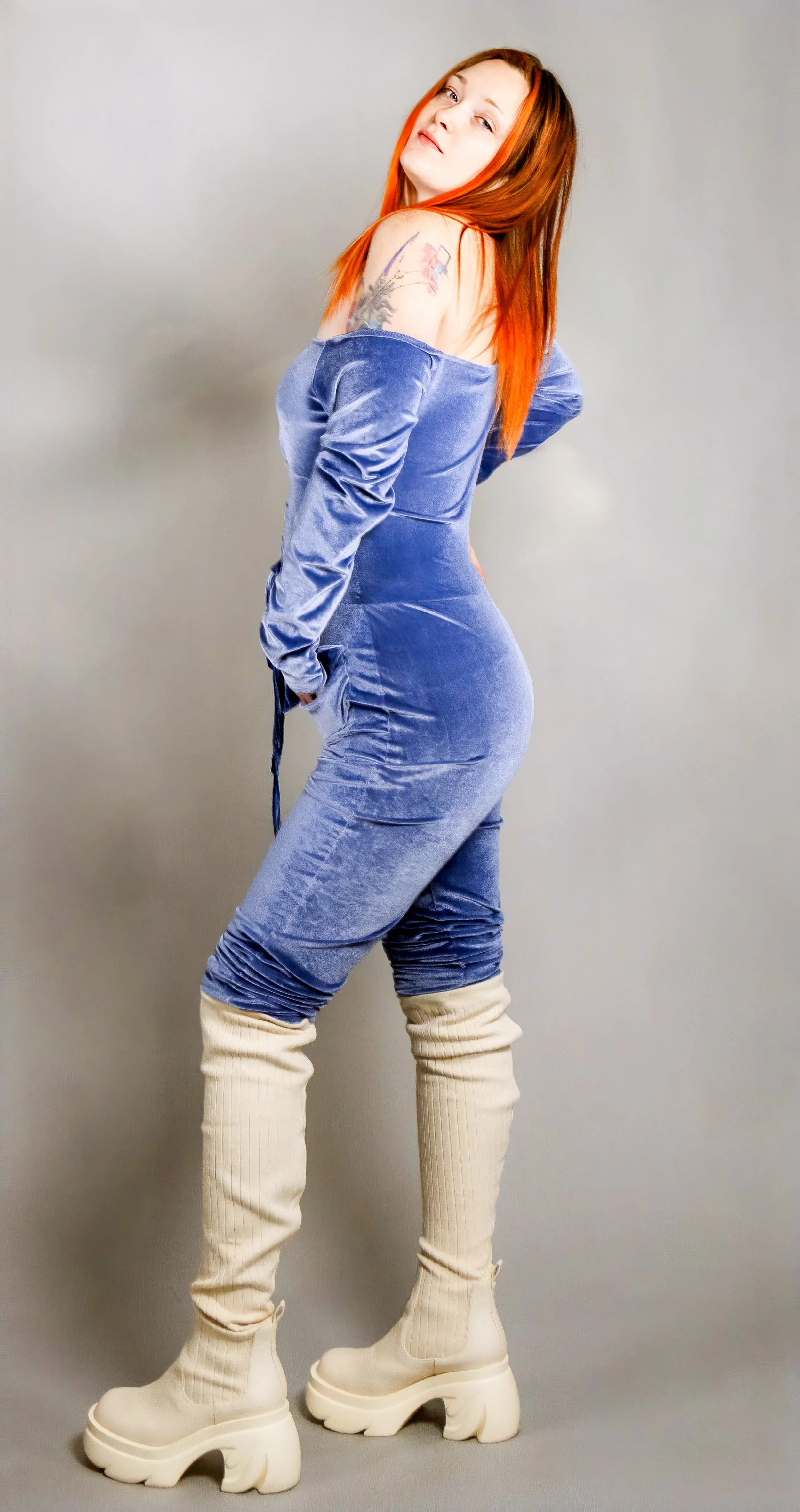 Woman with long red hair wearing a blue velvet off-shoulder jumpsuit, cream-colored over-the-knee boots, posing against a plain gray background.